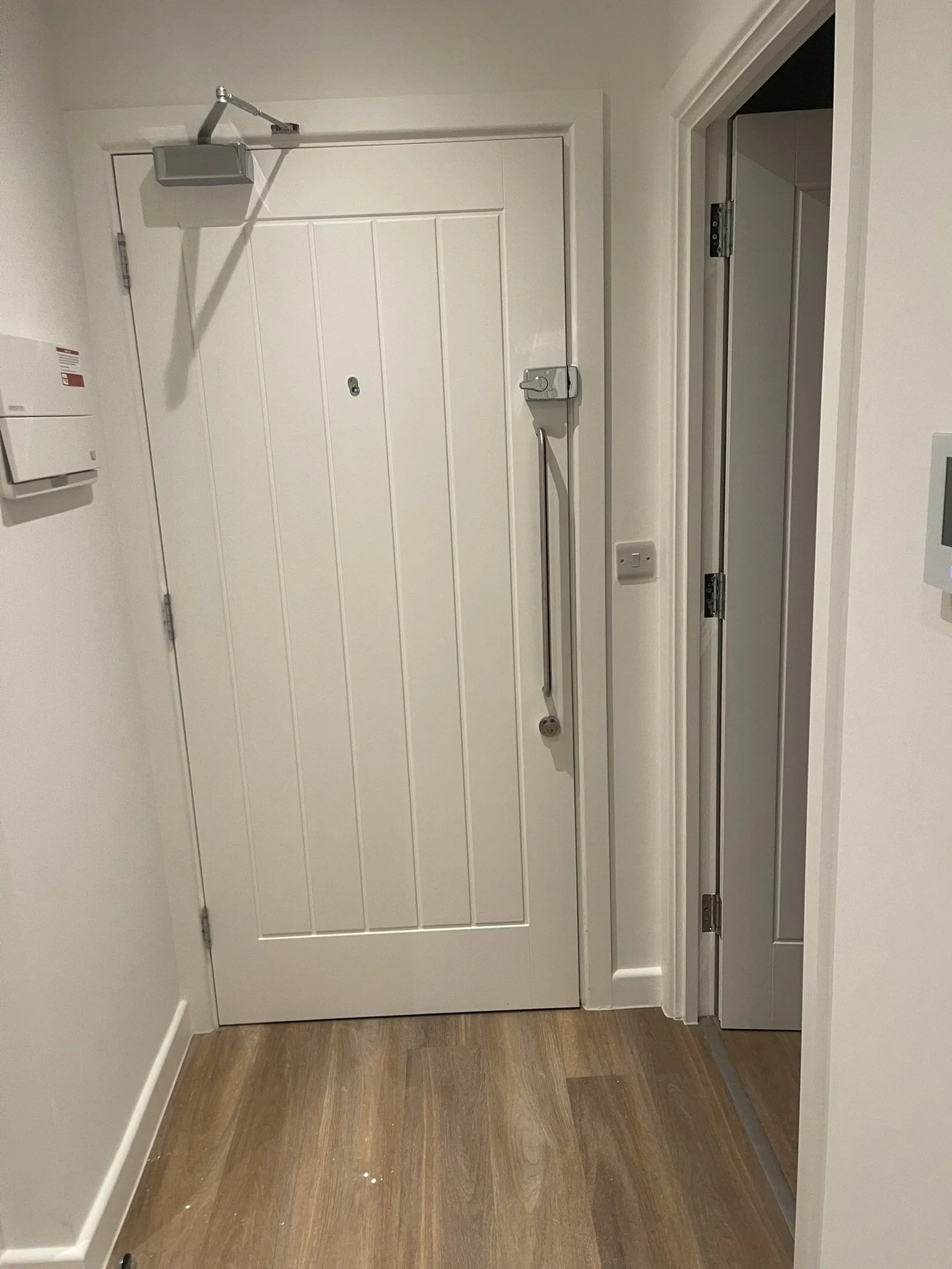 White paneled entry door with overhead door closer, security latch, and adjacent open interior doorway in a hallway.