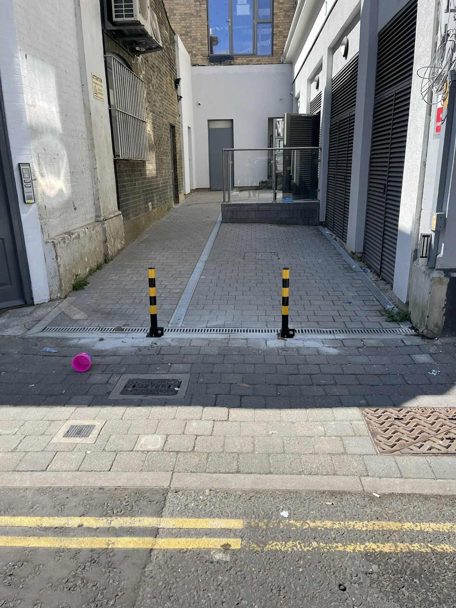 Paved alley entrance with black-and-yellow bollards, security gates, brick walls, and building doors.