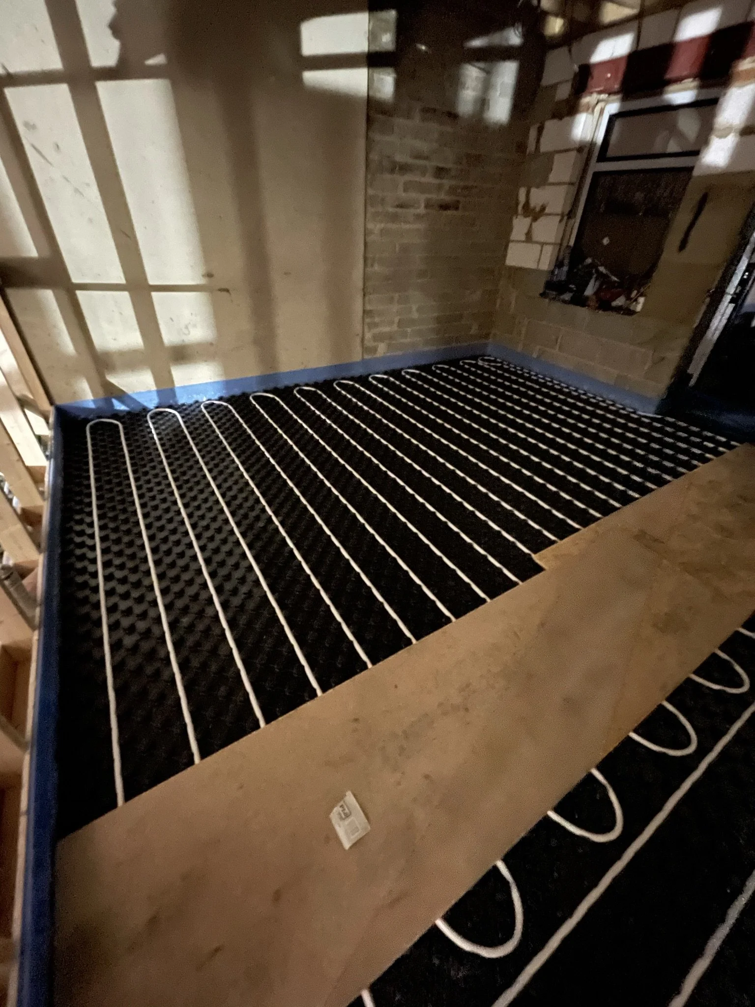 Electric underfloor heating cables laid across a black insulated floor beneath wooden boards in a brick-walled room.