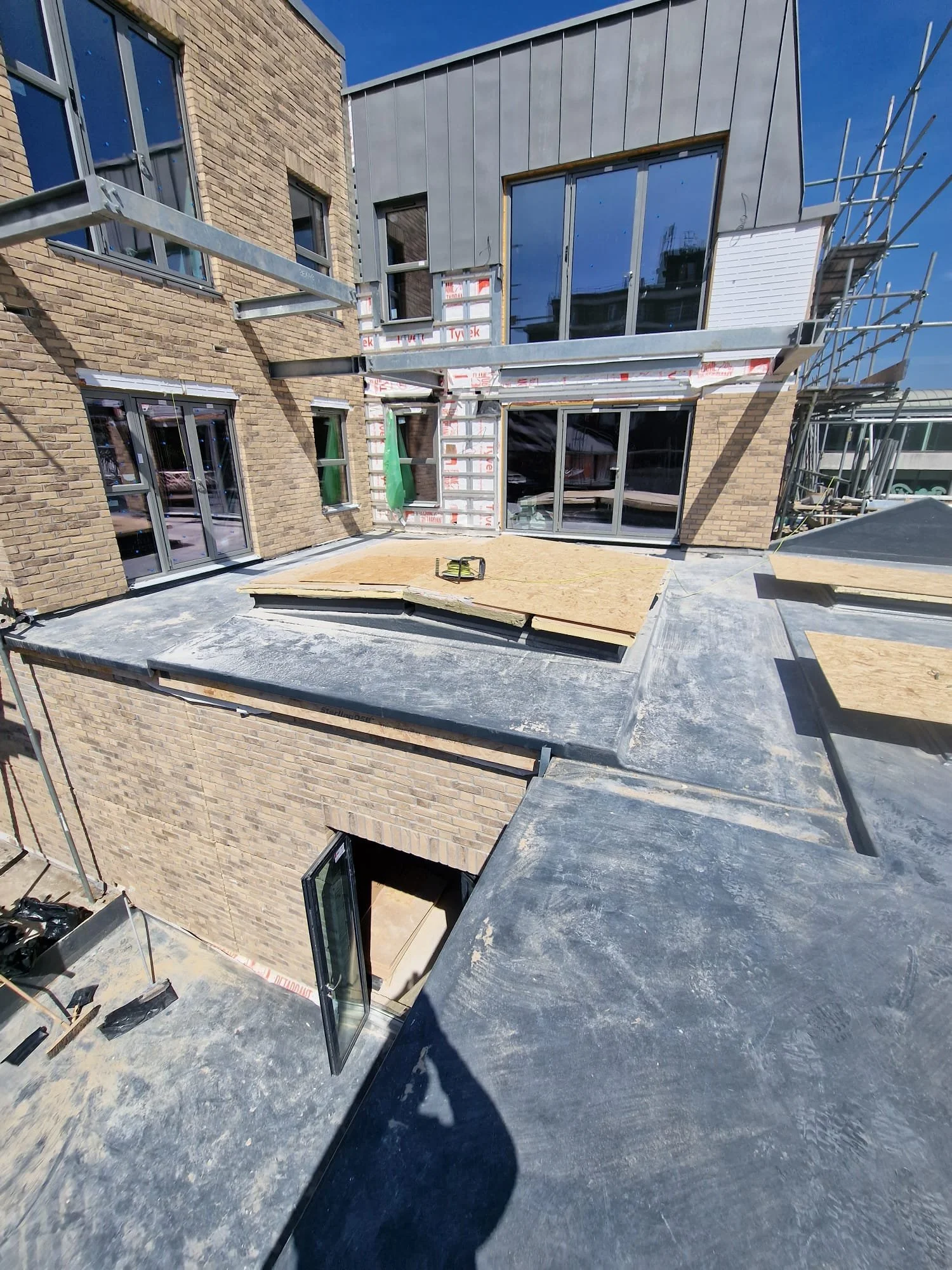 Modern brick building under construction with large windows, metal cladding, scaffolding, and unfinished rooftop terraces under a clear blue sky