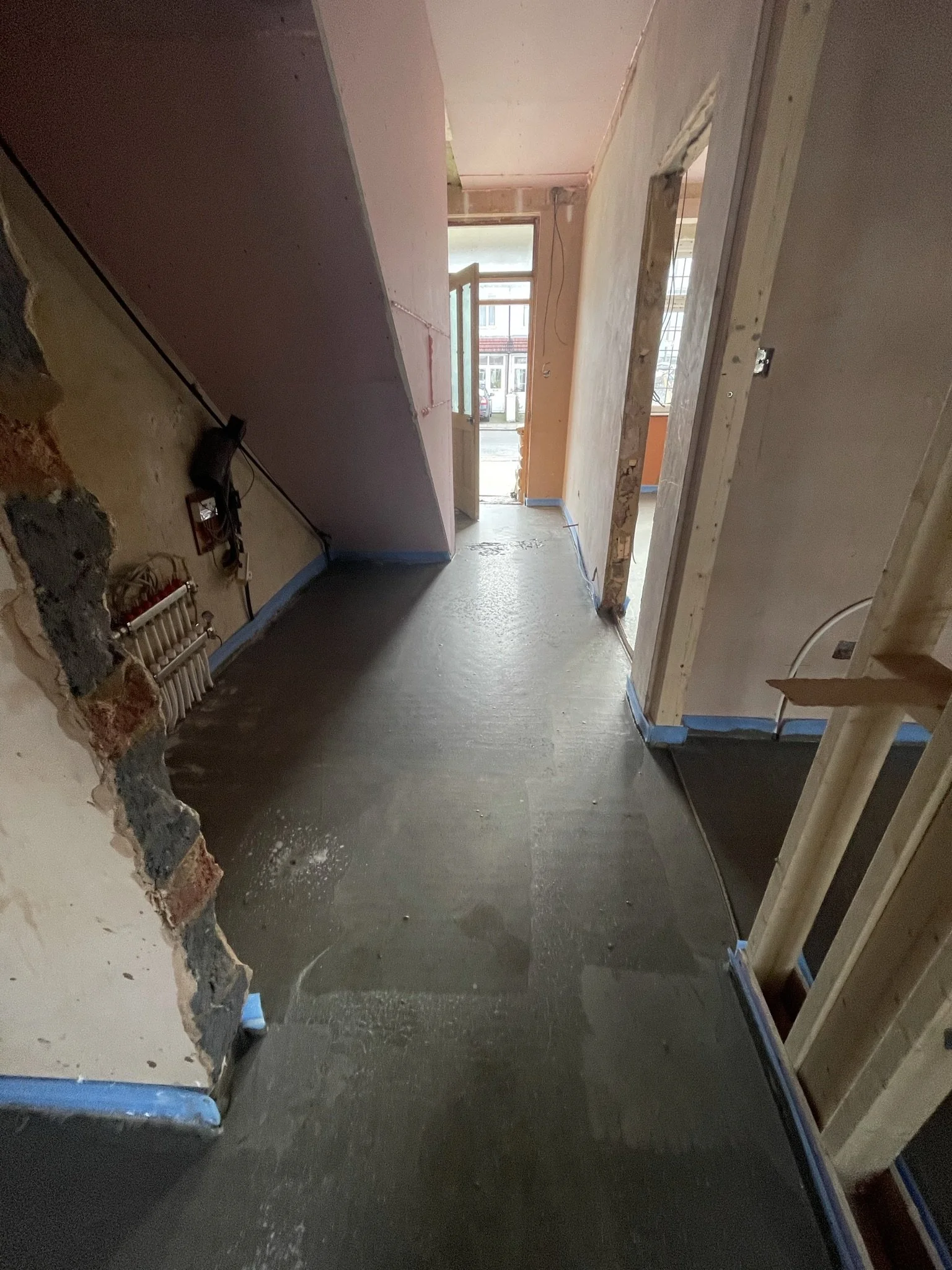 Partially renovated home hallway with exposed brick walls, unfinished doorways, gray concrete flooring, blue painter’s tape, and a street-facing entrance.