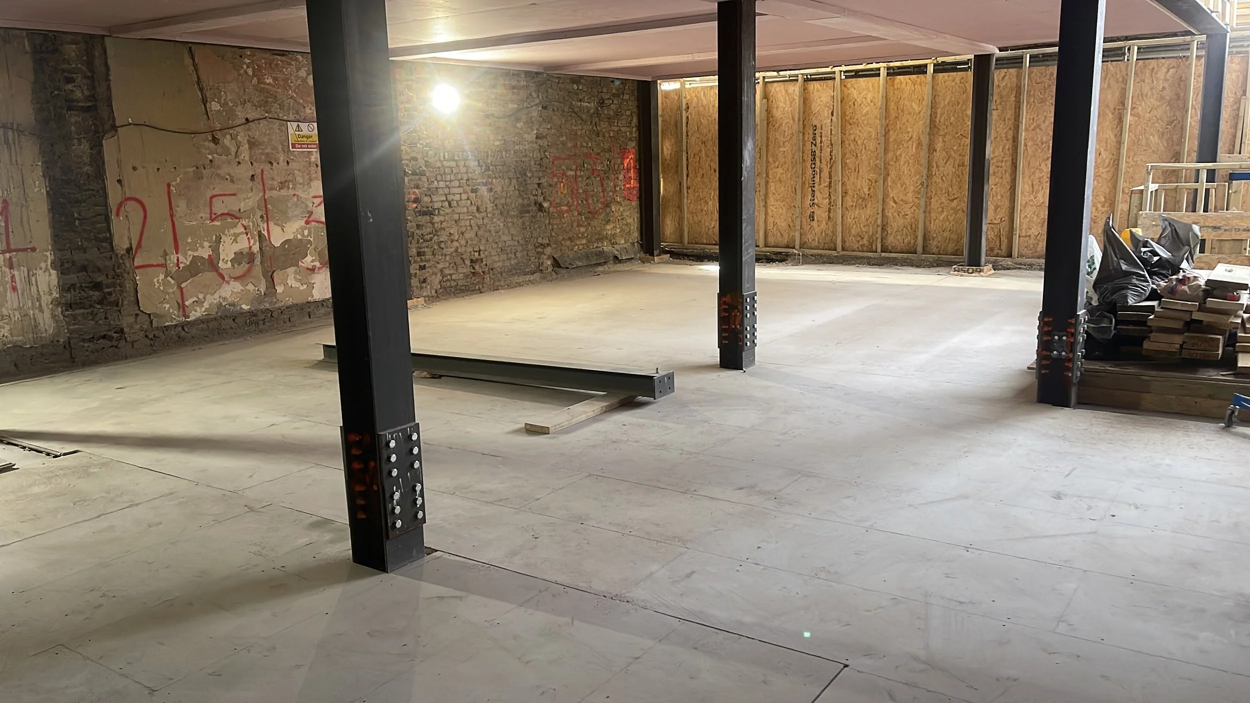 Renovated basement with exposed brick walls, black steel support columns, concrete floor, plywood wall framing, and construction materials.