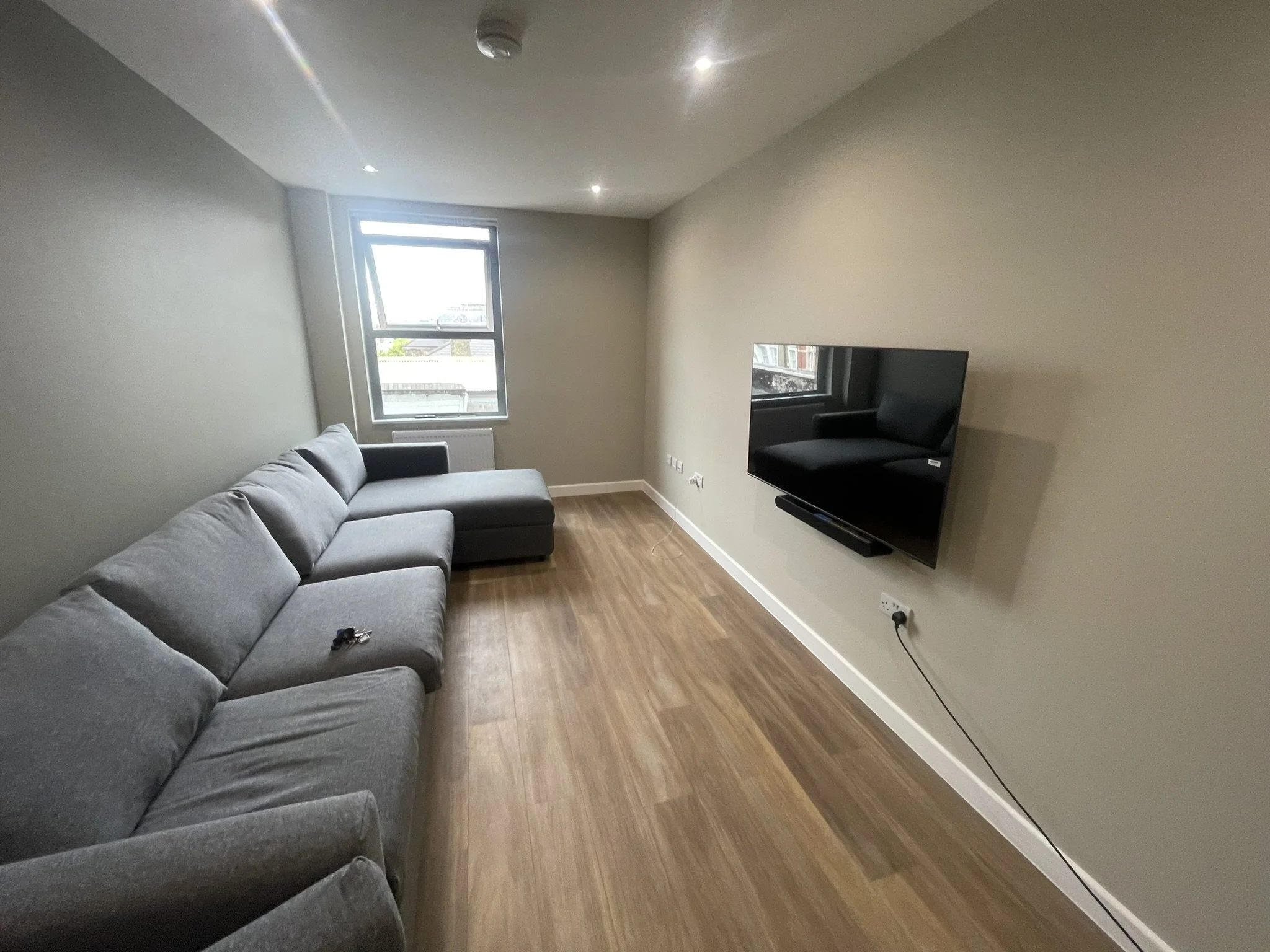 Modern living room with gray sectional sofa, wall-mounted flat-screen TV, wood-look flooring, window, and recessed lighting