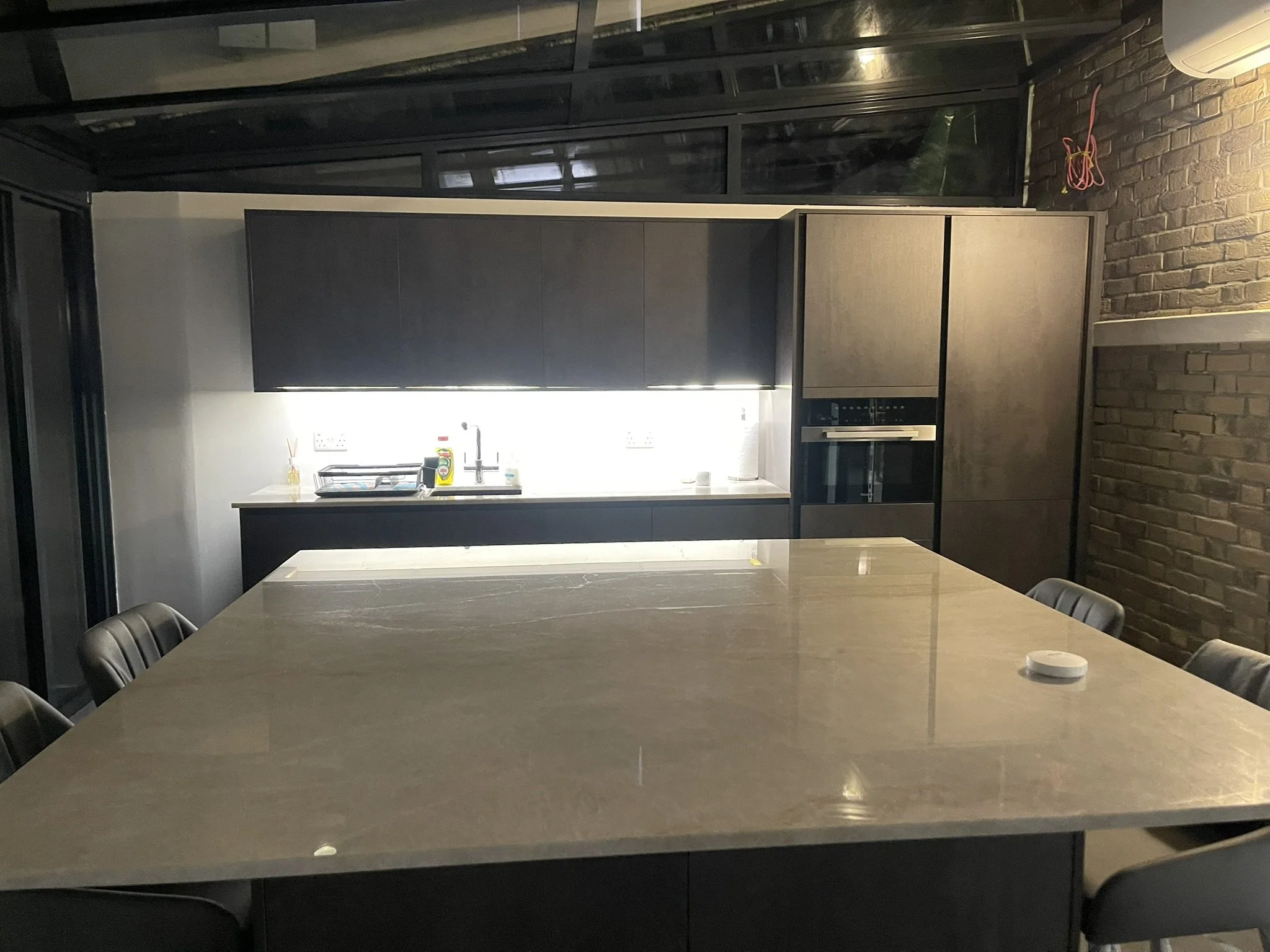 Modern dark kitchen with a large marble island, black cabinets, built-in appliances, bar stools, and exposed brick walls