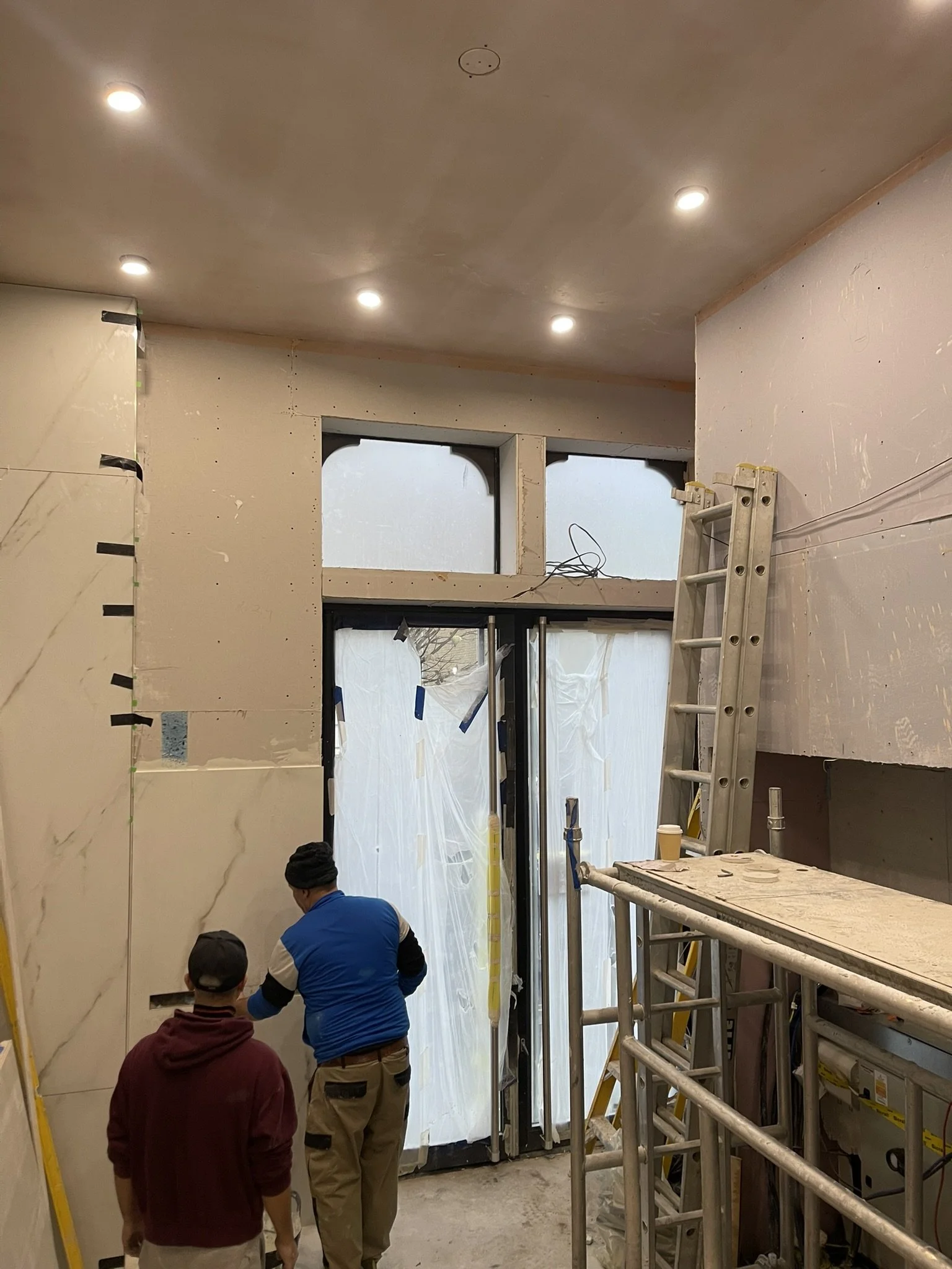 Construction workers renovating an interior with tiled walls, recessed ceiling lights, covered windows, and scaffolding