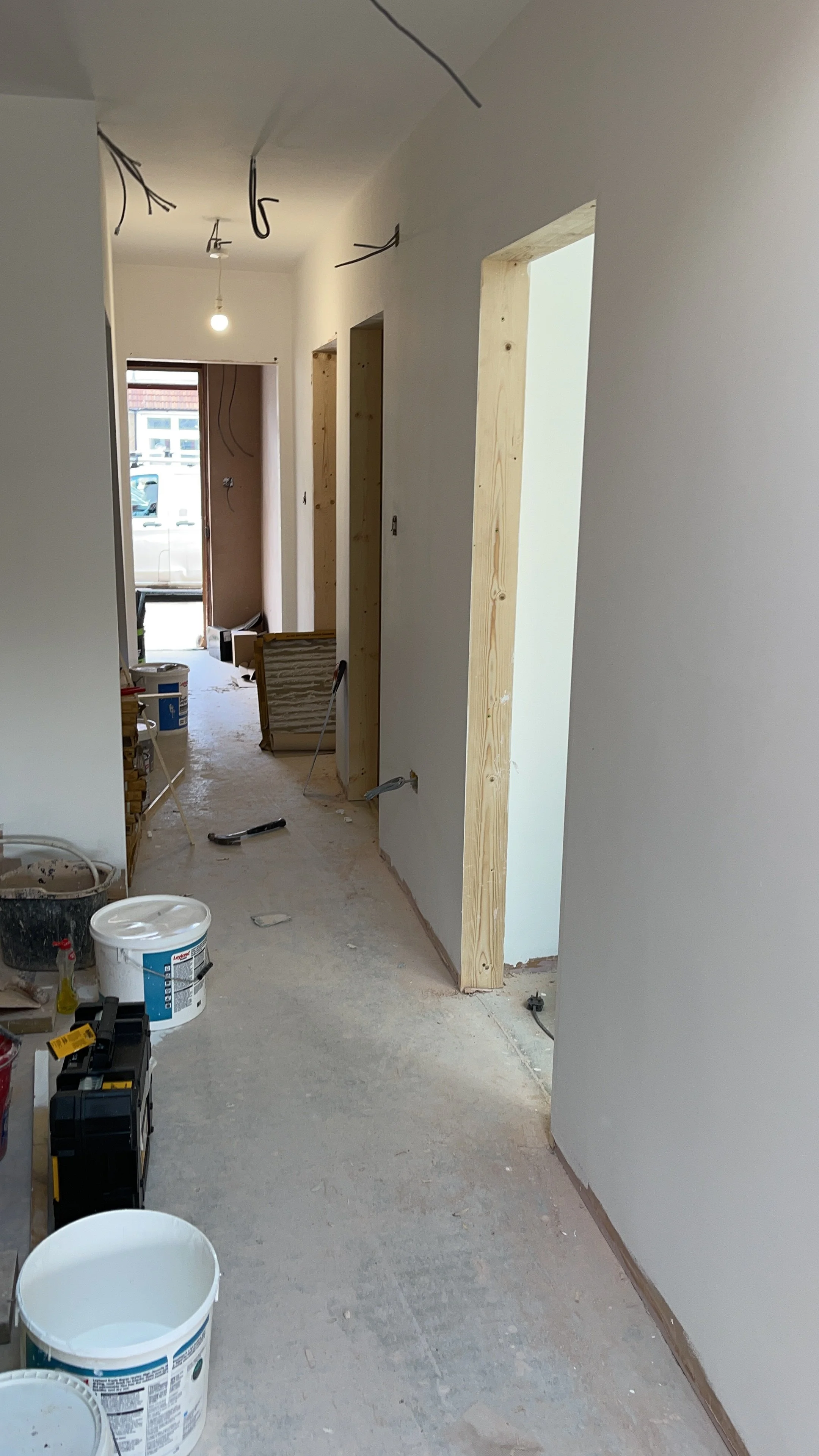 Interior hallway under construction with unfinished doorways, exposed wiring, paint buckets, tools, and drywall.