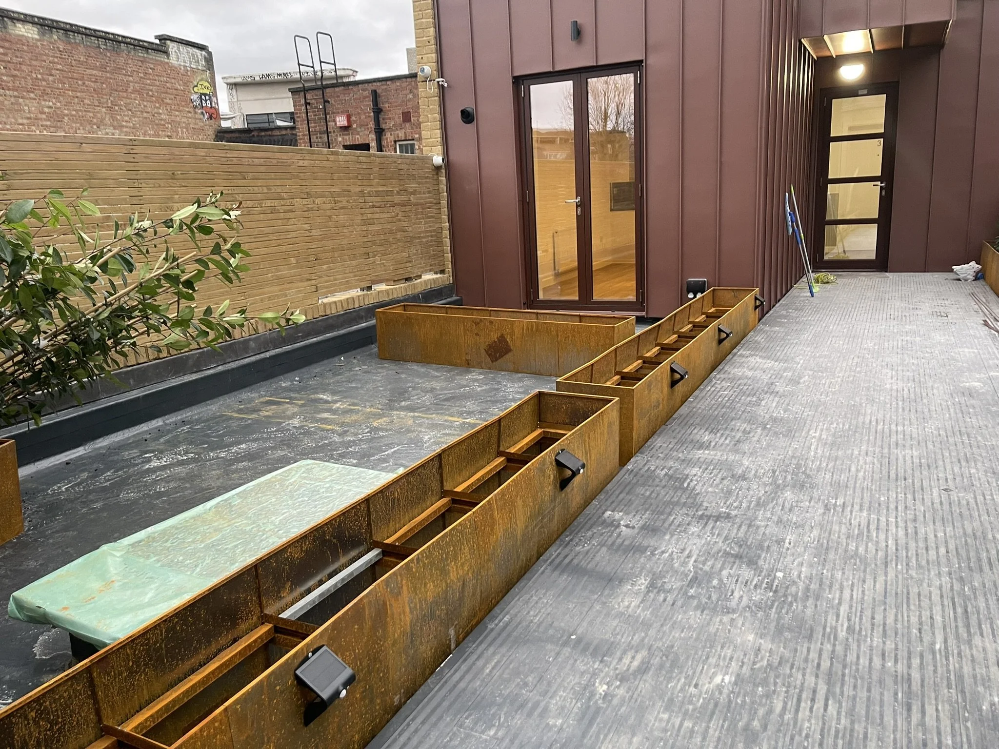 Rooftop terrace with rusted steel planters, glass doors, brick walls, and cleaning tools