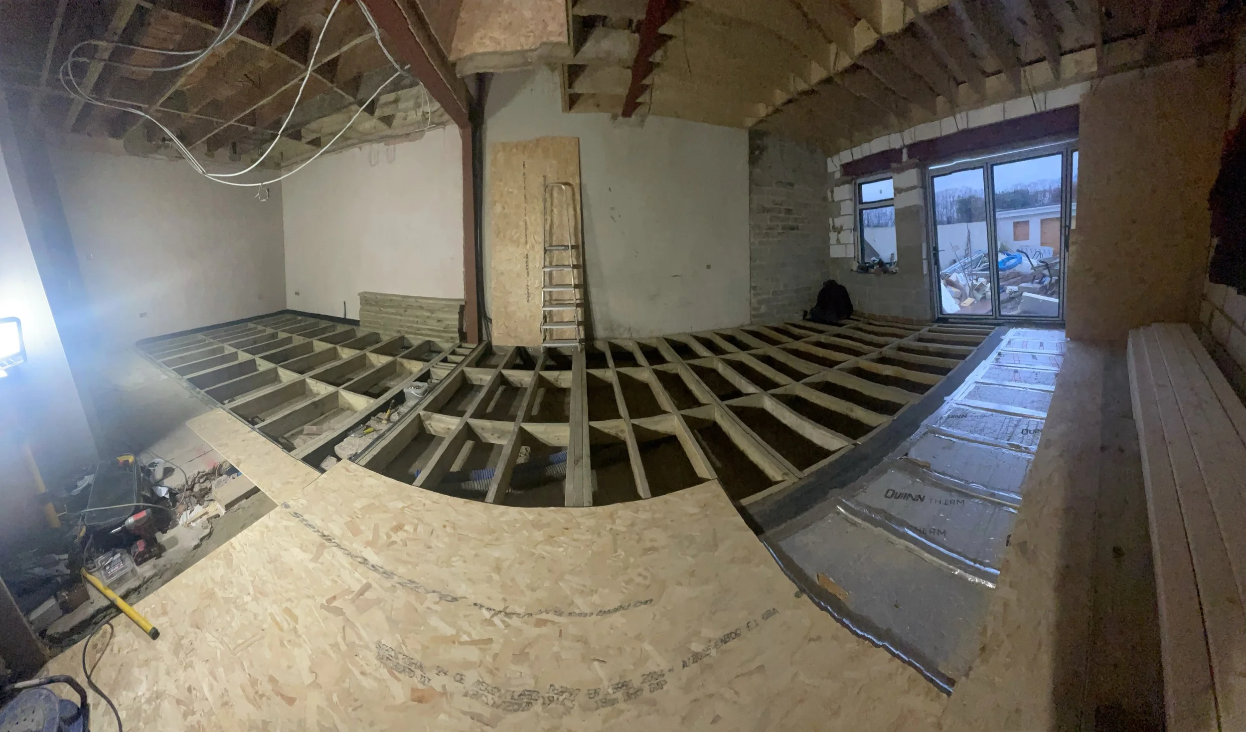 Interior of a house under renovation with exposed floor joists, plywood subflooring, insulation, construction tools, and unfinished walls