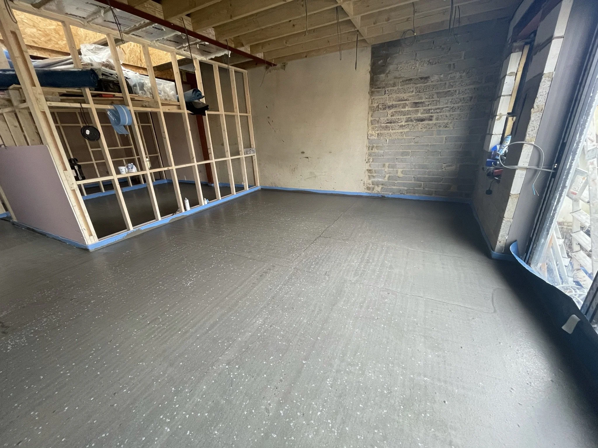 Partially renovated basement with exposed wall framing, brick wall, unfinished ceiling, storage shelves, and freshly poured concrete floor.