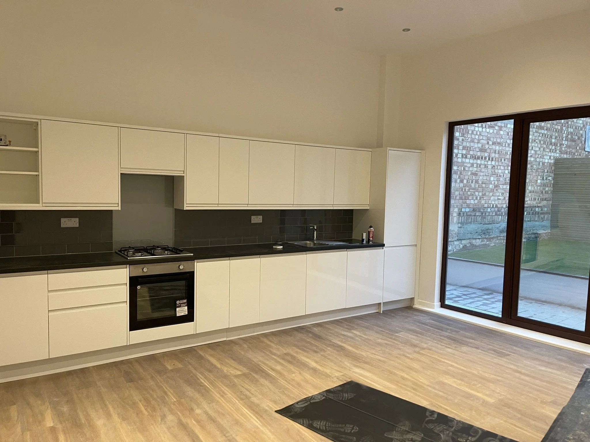Modern white kitchen with black countertops, gas cooktop, built-in oven, sink, dark tile backsplash, wood-look flooring, and large glass patio doors.