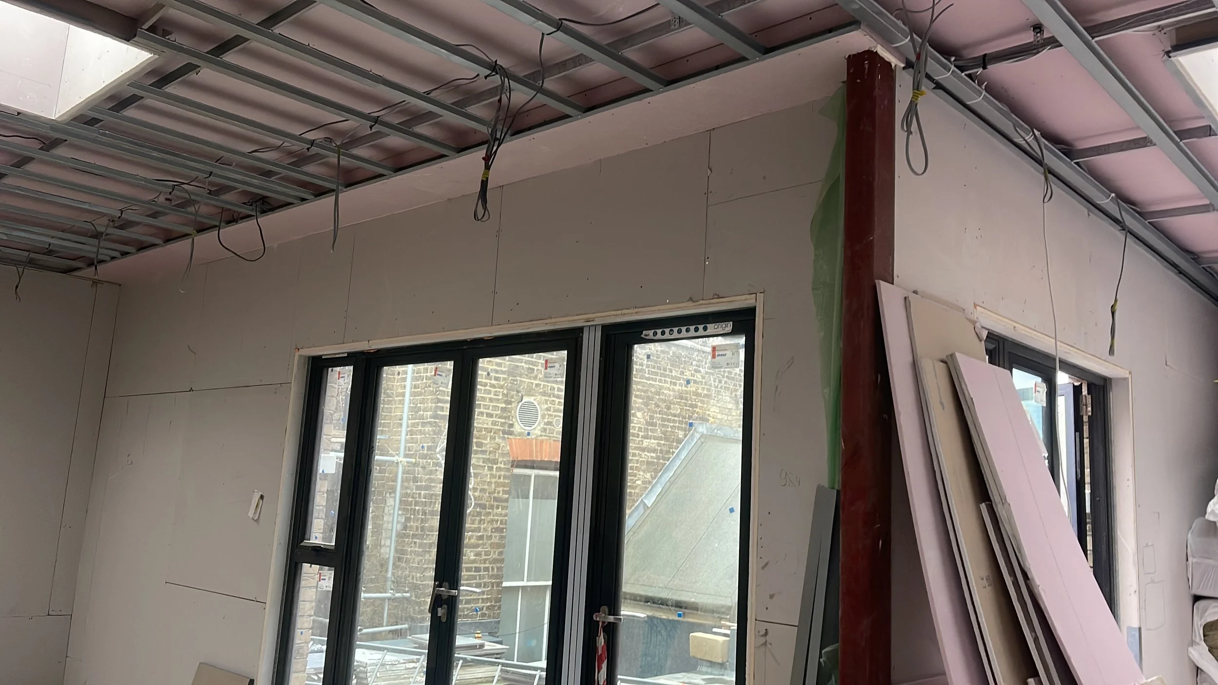 Interior construction site with unfinished drywall walls, exposed ceiling framing and electrical wires, black-framed windows, and stacked insulation boards.