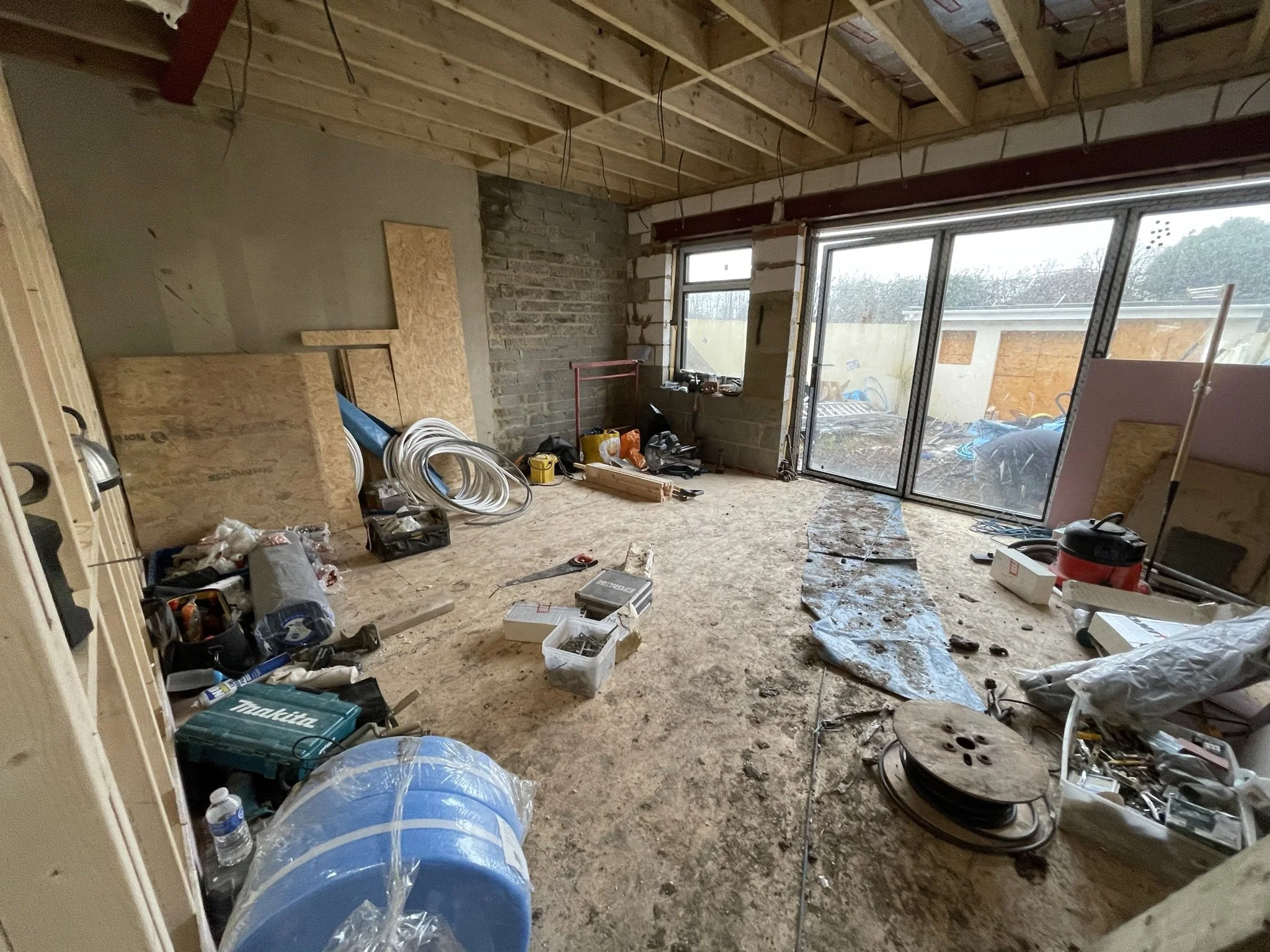 Unfinished room under renovation with exposed ceiling joists, construction materials, tools, coiled tubing, and large windows.