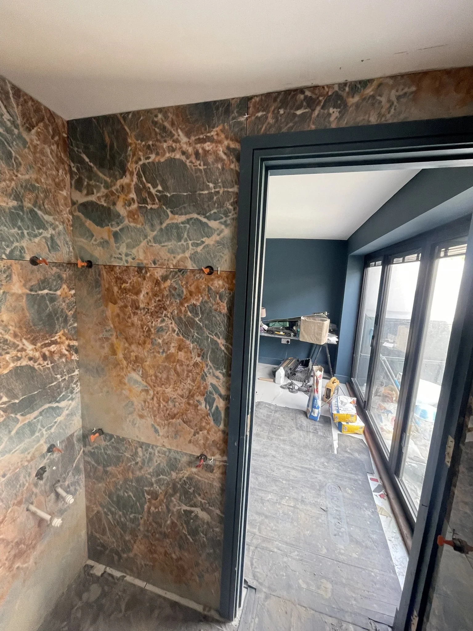 Bathroom under construction with striking brown-and-gray marble wall tiles, plumbing fixtures, and a dark-framed glass door.