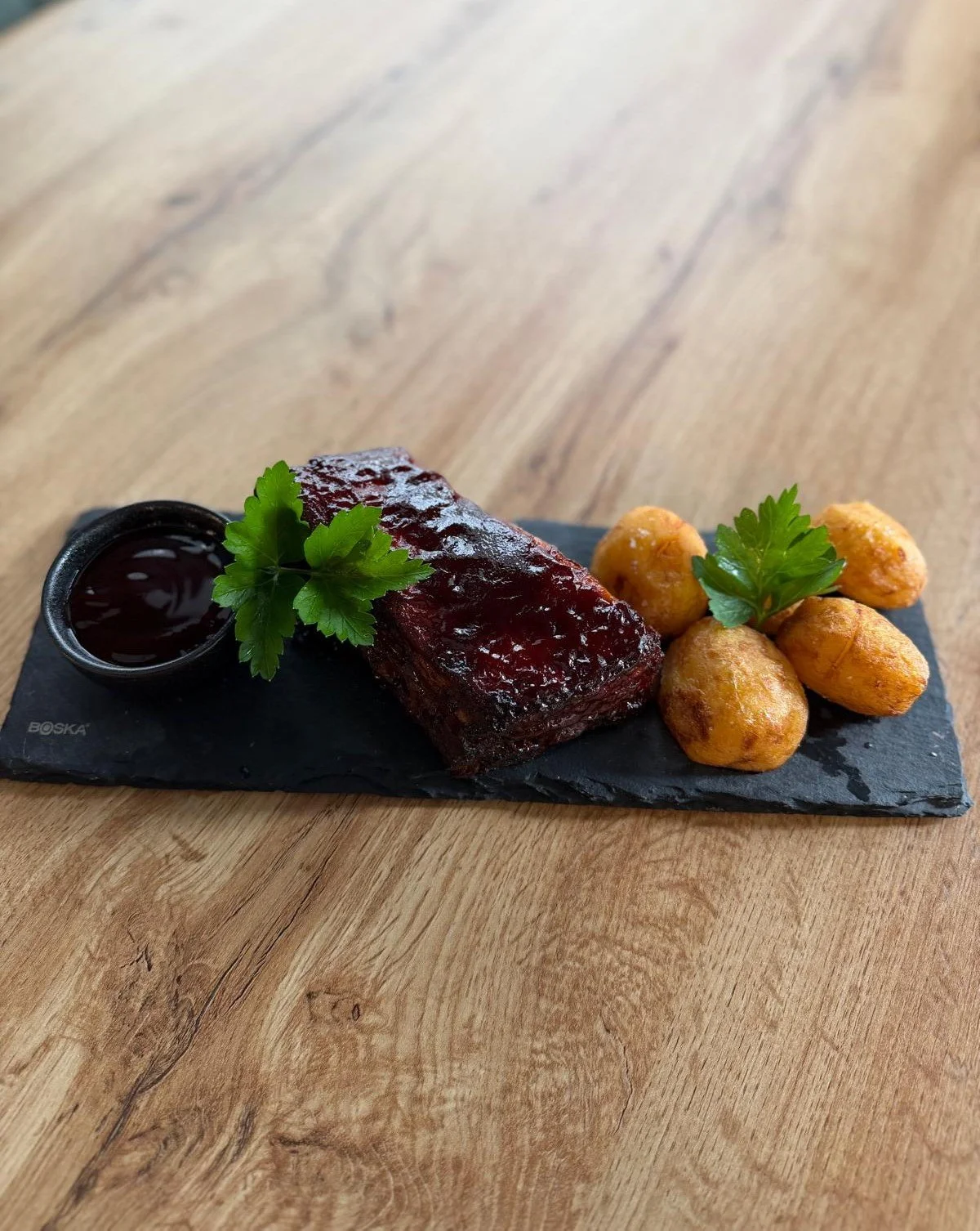 A serving of glazed beef short ribs garnished with parsley, served with crispy potato bites and a side of dark sauce on a black slate platter.