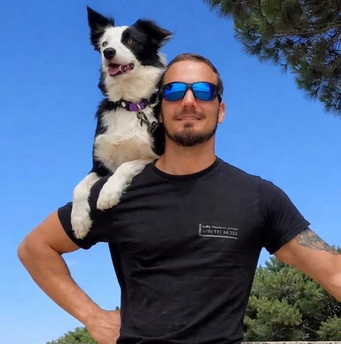 Un homme portant des lunettes de soleil bleues pose avec un chien noir et blanc sur son épaule, dans un environnement extérieur avec un ciel bleu et des arbres.