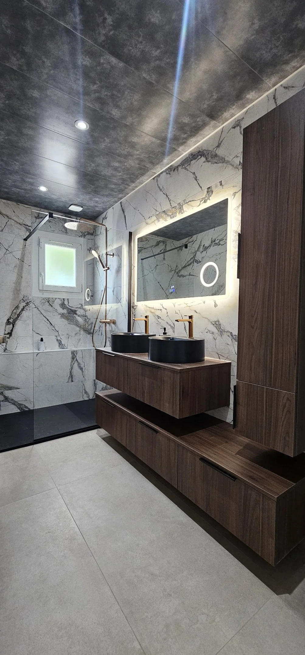 Salle de bain moderne avec deux lavabos noirs sur une surface en bois, miroir avec éclairage LED, murs en marbre blanc avec des veines noires, douche avec paroi en verre, fenêtre à l'arrière, plafond en béton ciré.