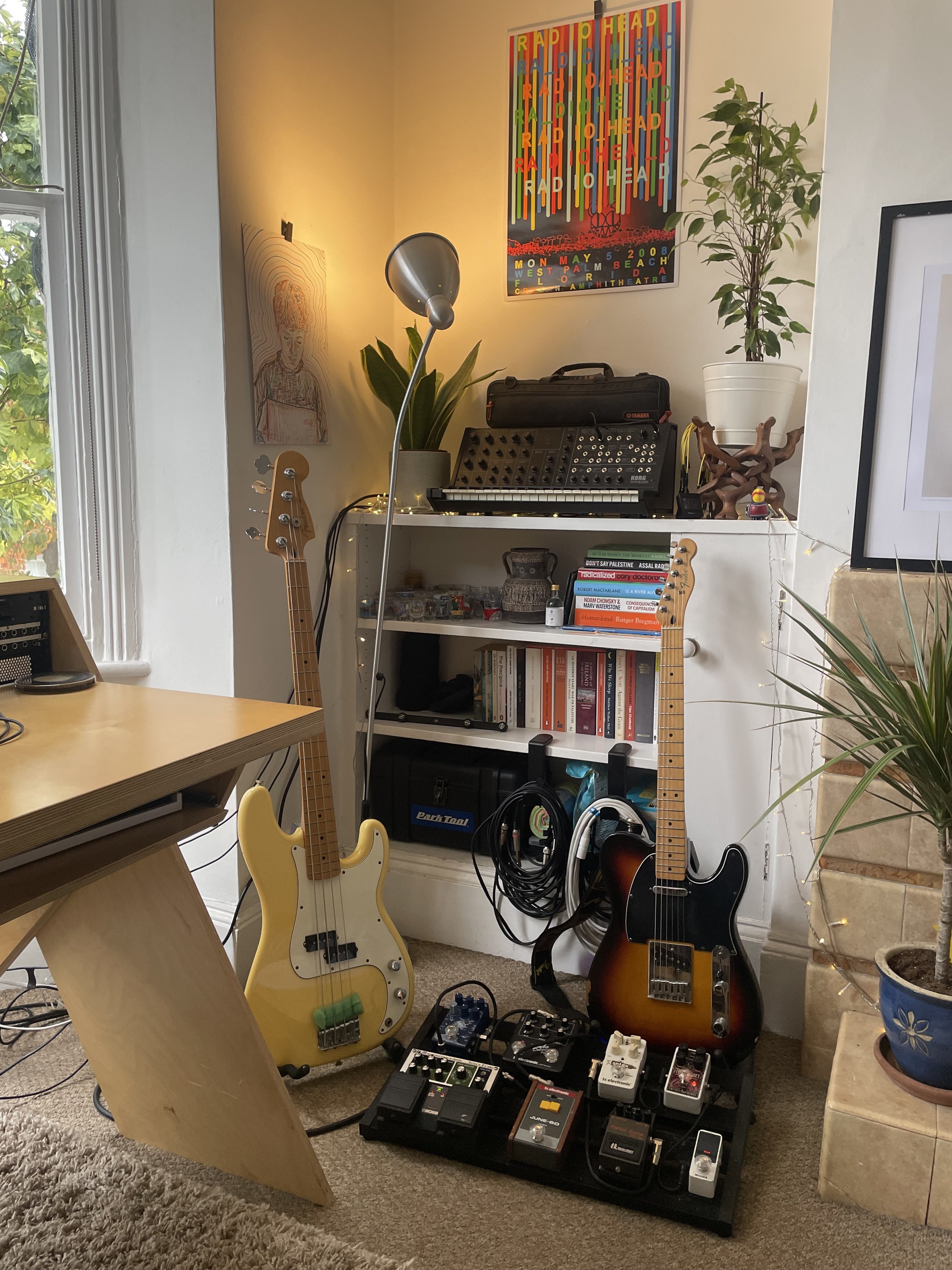 A room with musical instruments and related equipment. There are two electric guitars, one yellow and one sunburst, a synthesizer, and a pedalboard with multiple effects pedals. A bookshelf with books and decorative items is against the wall, with a poster titled 'Radiohead' hanging above it. There is a potted plant and a framed artwork on either side of the room, and a window with greenery outside.