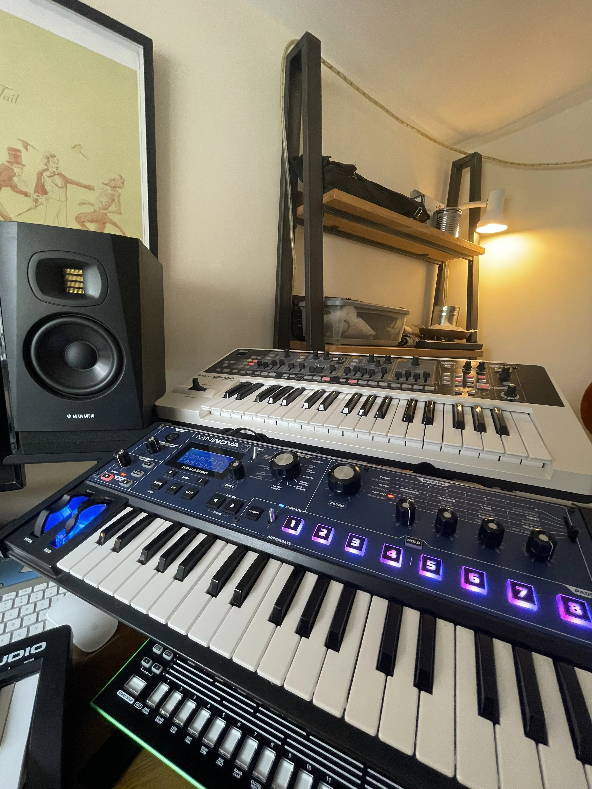 A music production setup featuring a keyboard synthesizer with illuminated purple buttons, a black studio monitor speaker, and a smaller MIDI controller with a green LED light. In the background, there's a wooden shelf with various items, including a black bag, a framed picture, and metal cans. A lamp on the wall provides warm lighting.