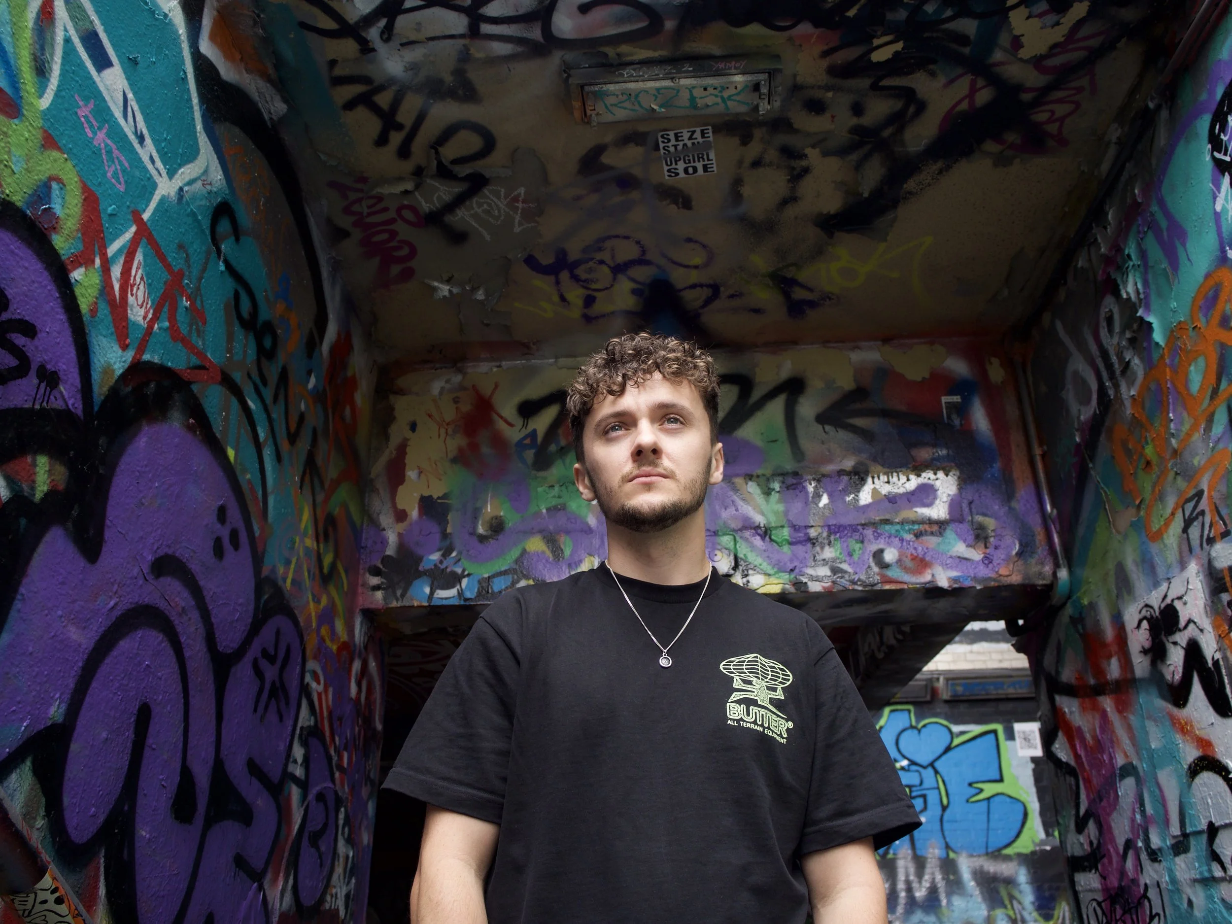 A young man with curly hair and a beard wearing a black t-shirt and silver necklace standing inside a graffiti-covered tunnel with colorful spray-painted walls.