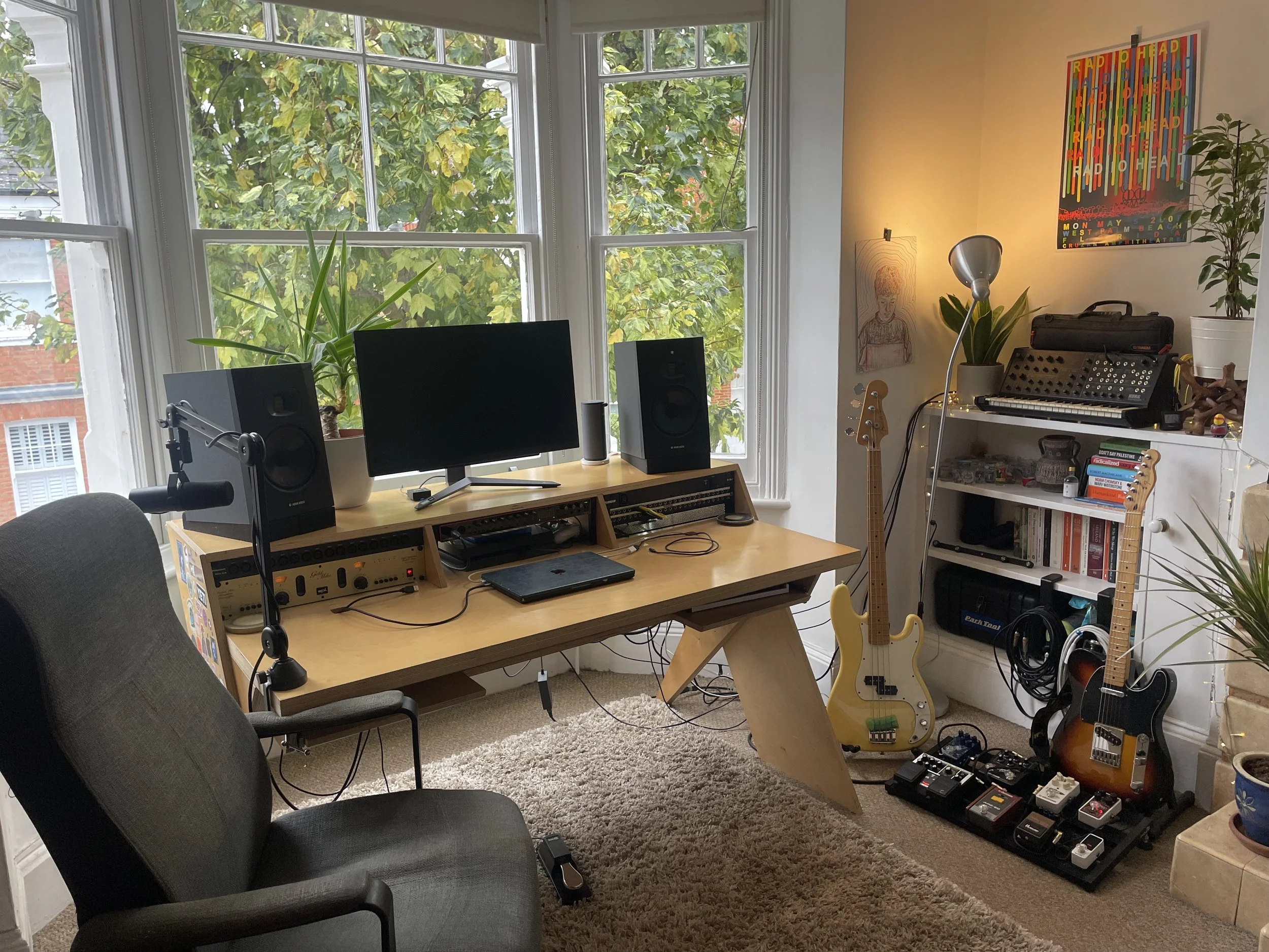 A home music studio with a computer monitor, studio speakers, and various musical instruments including an electric guitar, a ukulele, and a keyboard. There are plants on the window sill and a colorful RAYMOUNT poster on the wall. The room has a large window showing green trees outside.