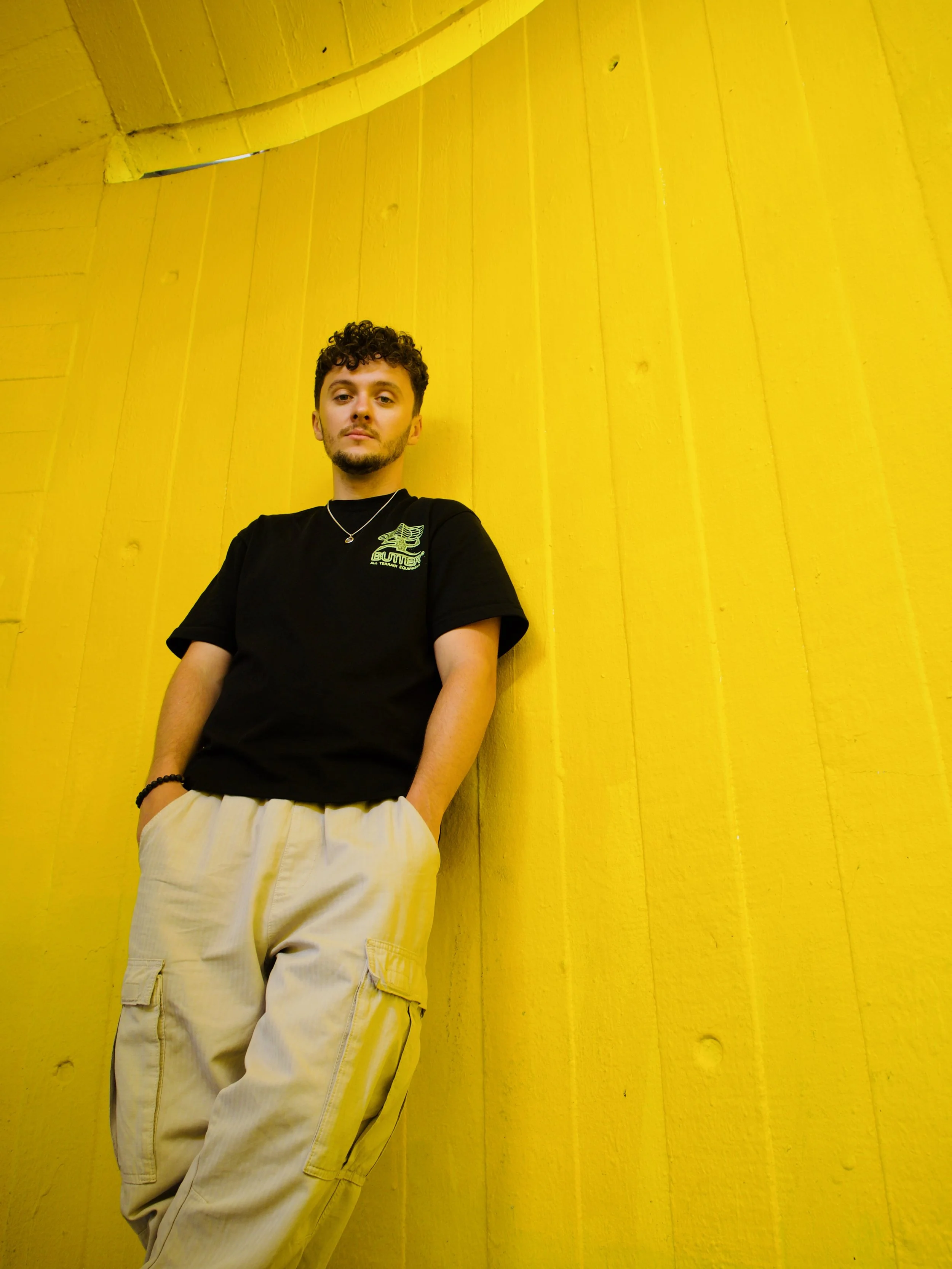 A young man with curly hair and a beard standing against a yellow wooden wall. He is wearing a black t-shirt with a logo on the chest, beige cargo pants, a necklace, and a bracelet on his left wrist.