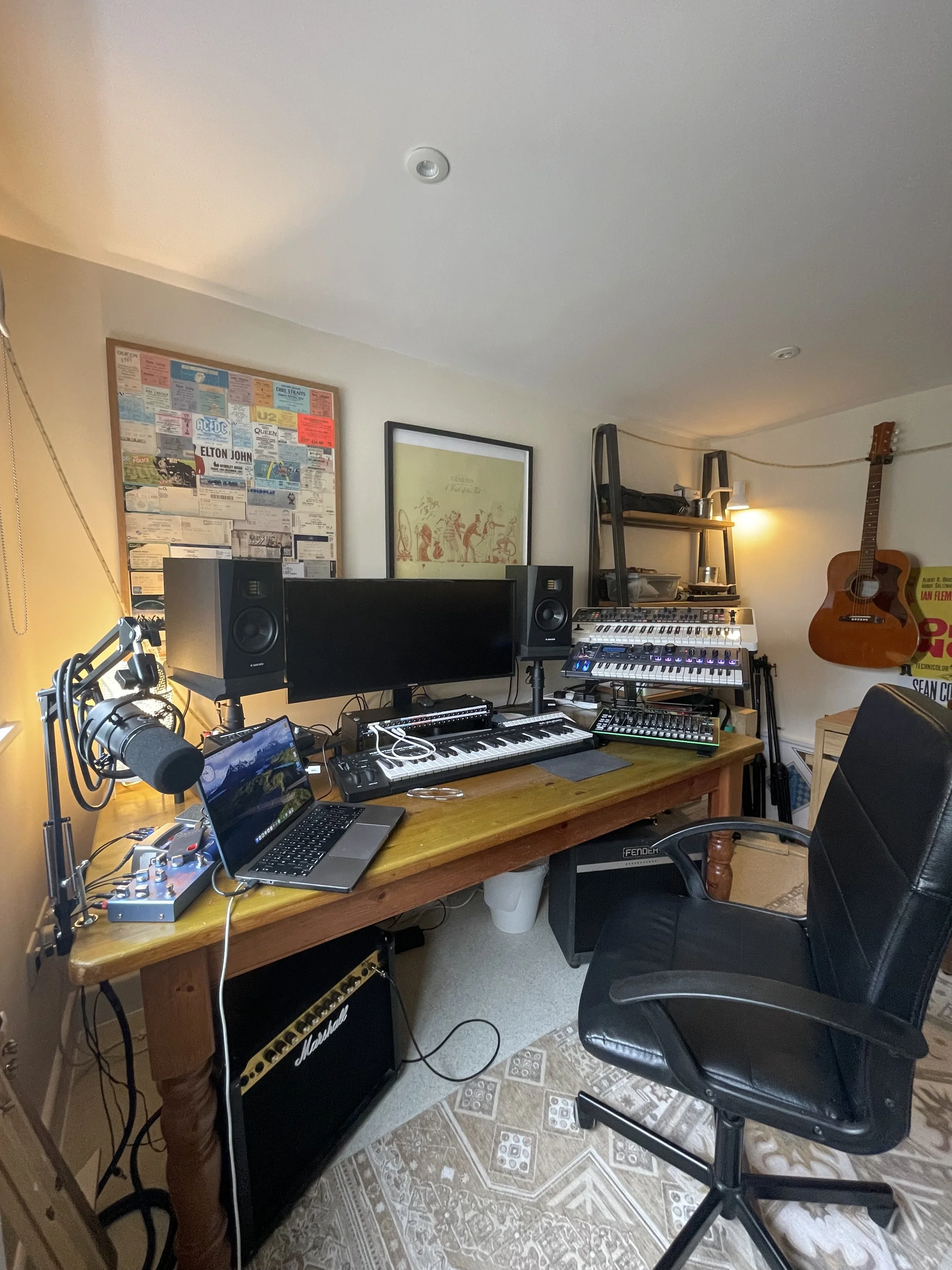 A home music studio with a wooden desk holding a laptop, a small audio interface, speakers, and electronic music equipment. The wall features posters, framed art, and a guitar hanging on the wall. A black office chair is positioned in front of the desk, and a larger speaker is on the floor. There is a shelving unit with various items nearby.