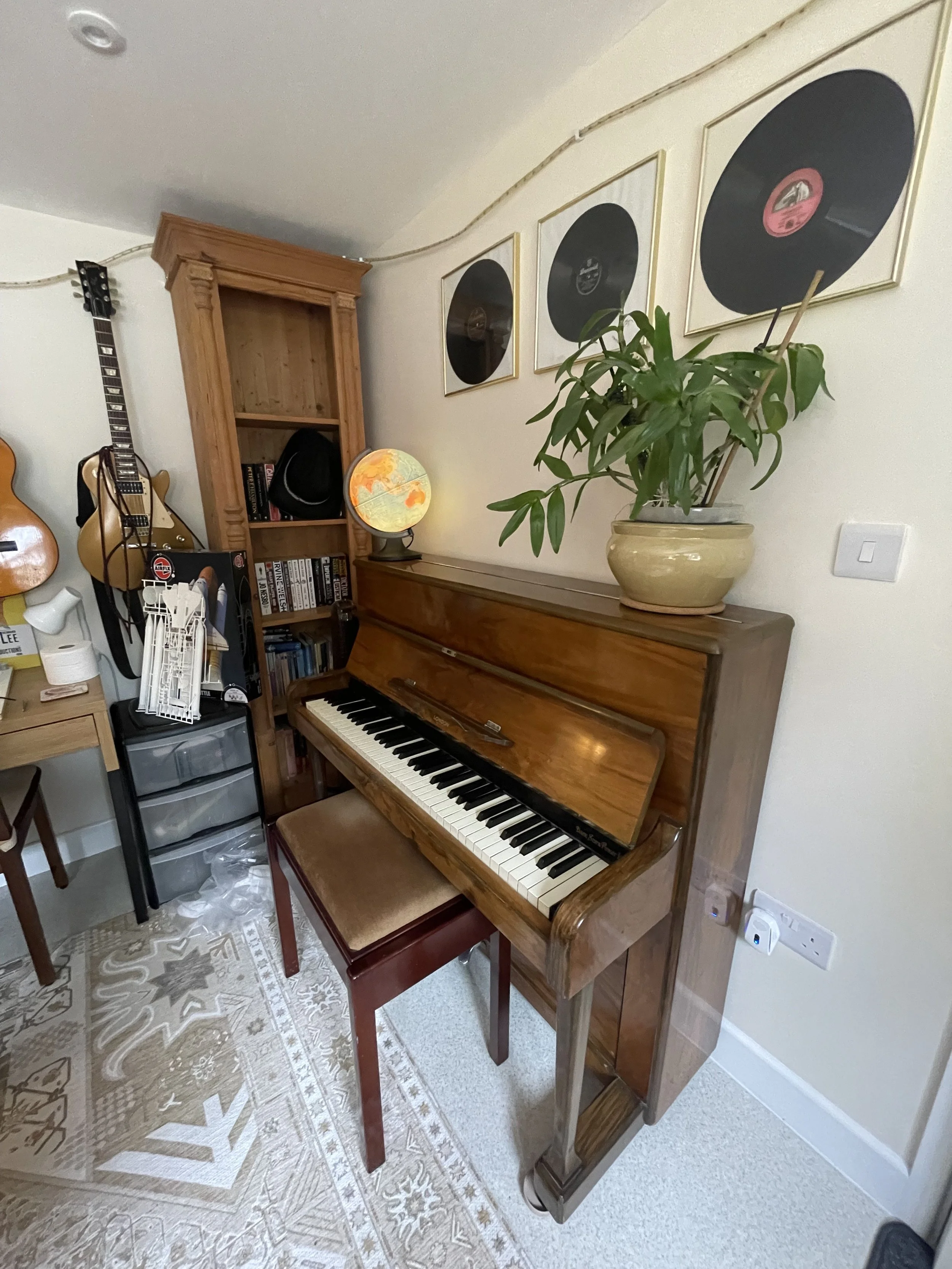 A room with a wooden upright piano, a potted plant on top, framed black vinyl record artwork on the wall, a guitar mounted on the wall, a small globe lamp, a bookshelf, and various household items. The floor has a patterned rug.