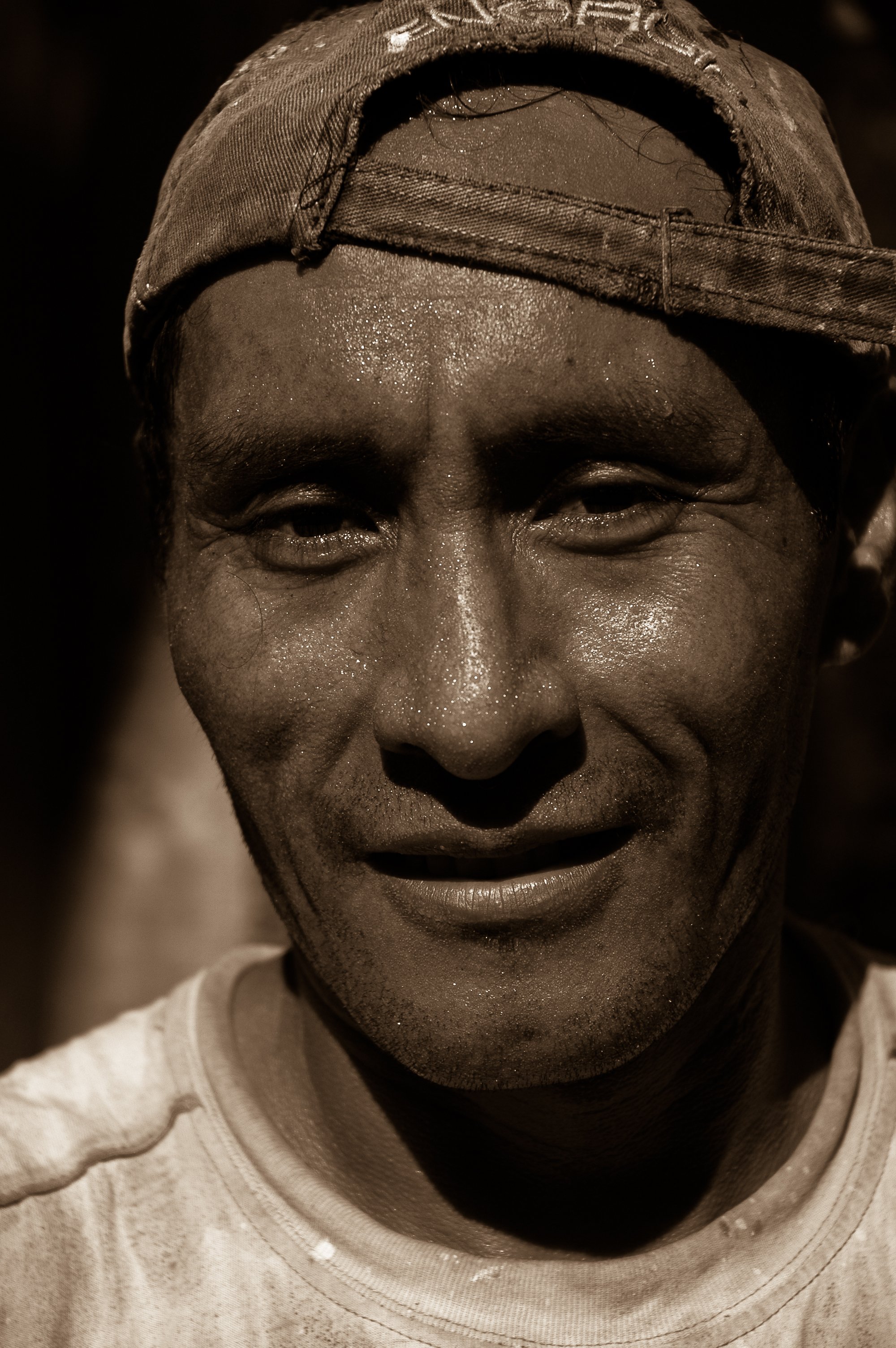 Close-up of a person's face wearing a backwards baseball cap. The person has wet, glistening skin, dark eyes, and a slight smile, with a plain light-colored shirt visible.