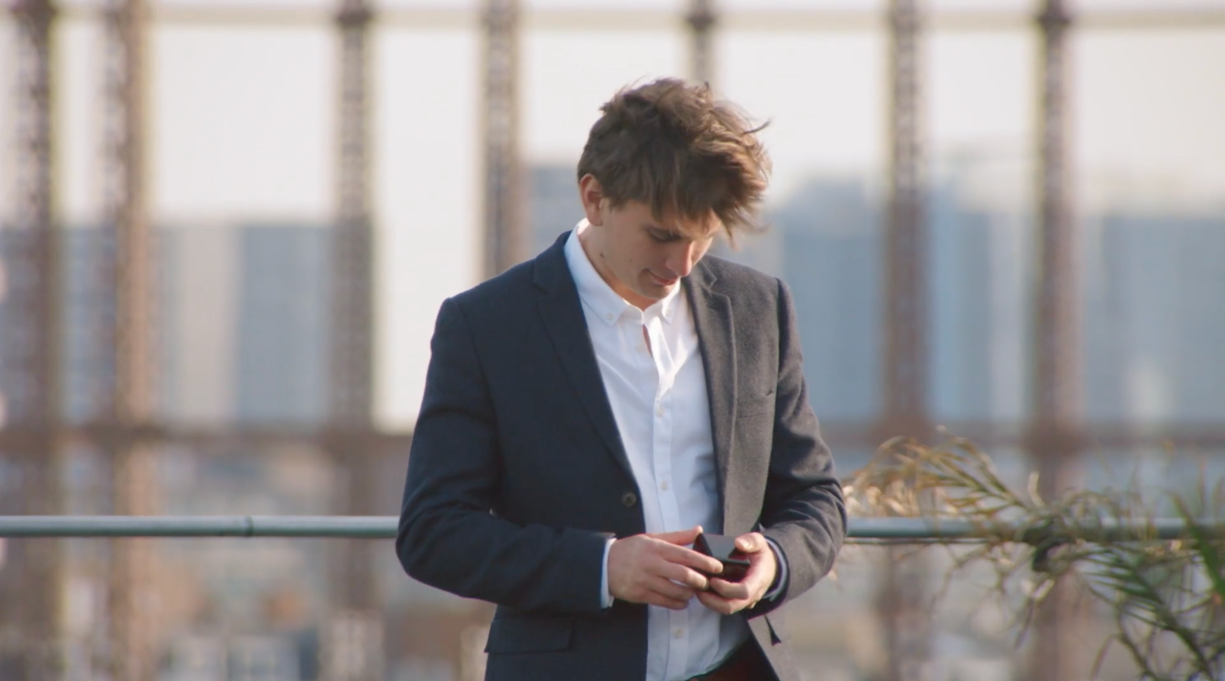 A young man in a suit looking down at his phone on a rooftop with a rail and some plants in the foreground.