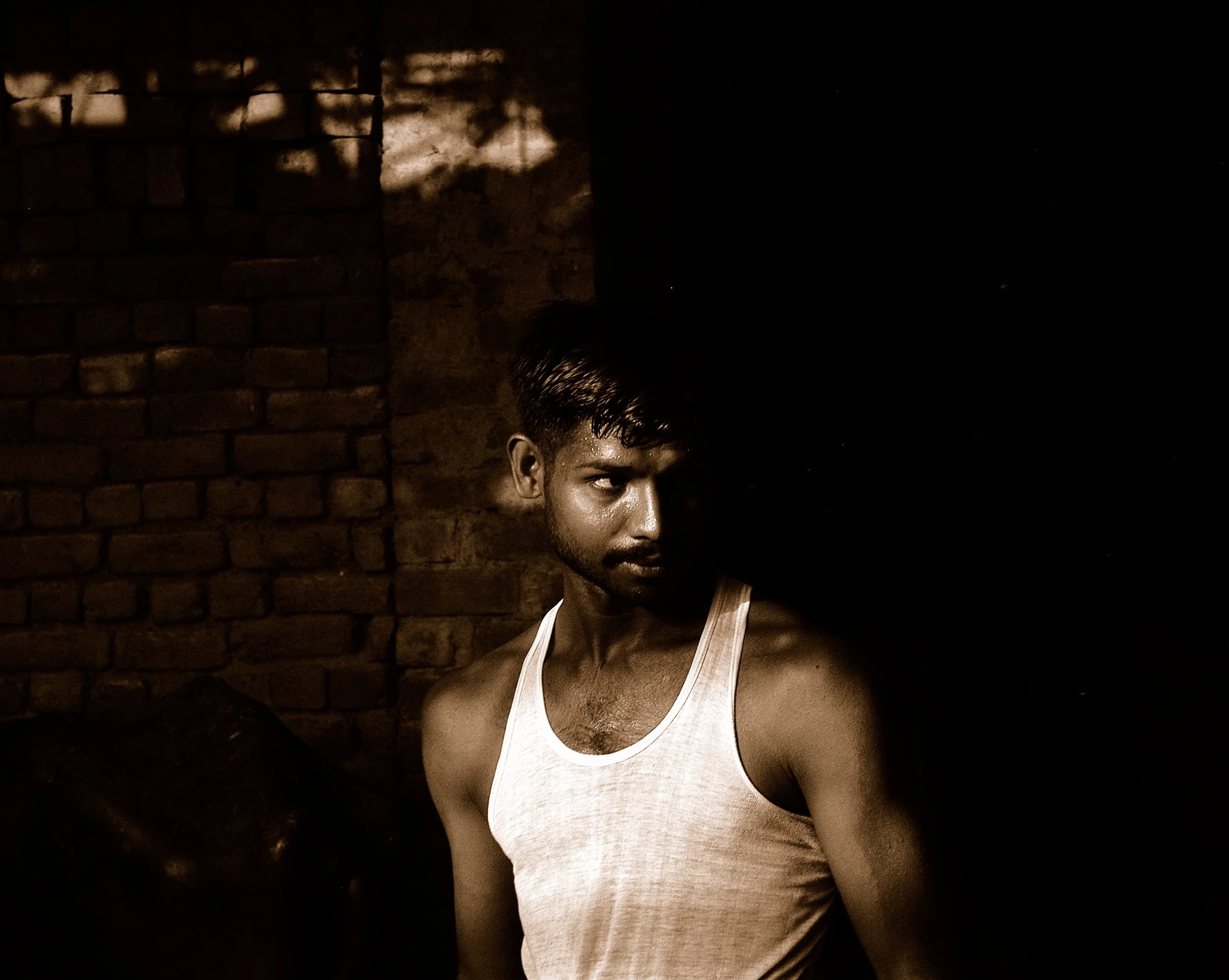 A young man with a muscular build and short, dark hair, wearing a white tank top, sitting against a brick wall at night with shadows cast on the wall.