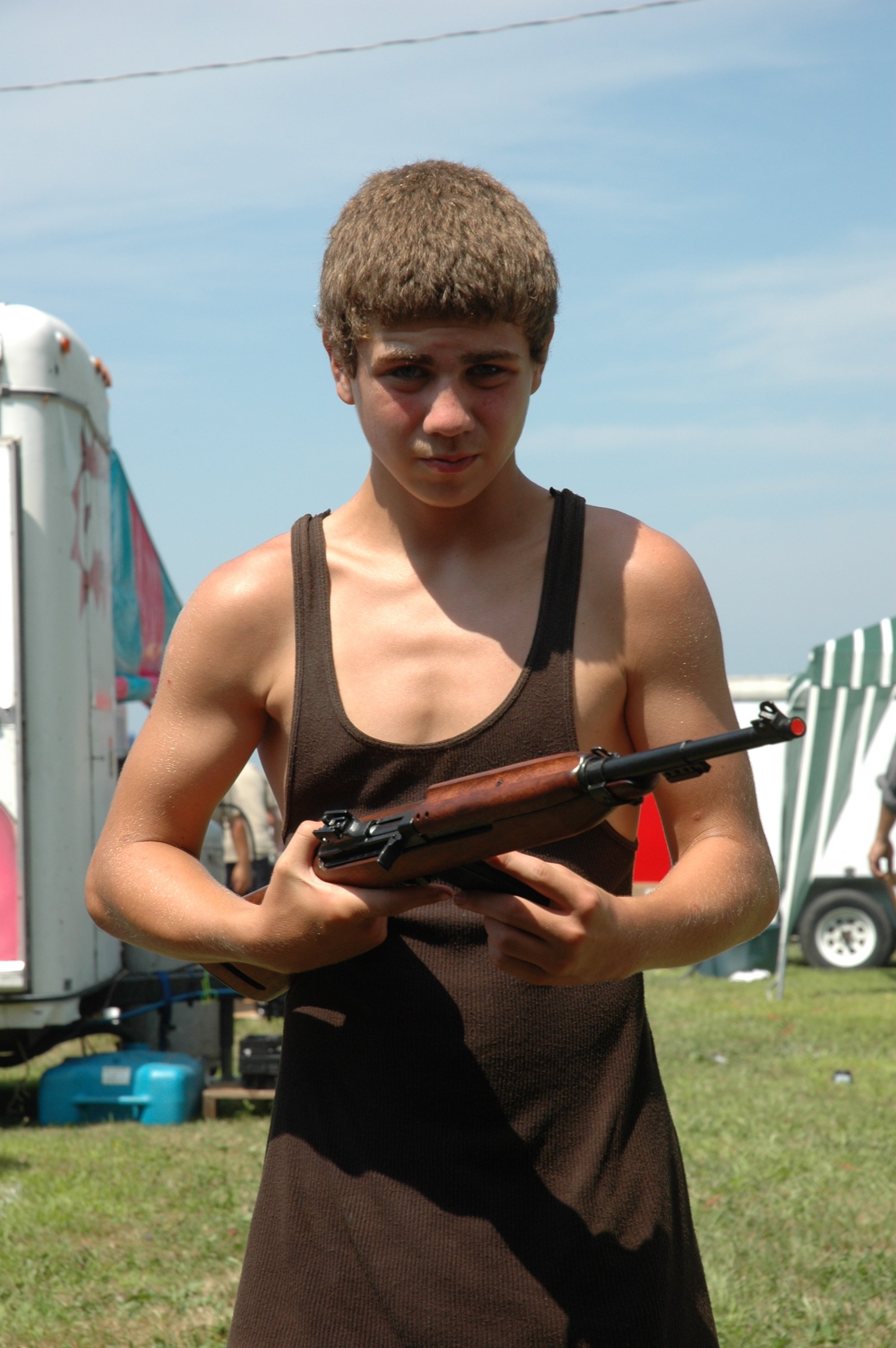 A young person with short brown hair wearing a black sleeveless top holding a firearm at an outdoor event with a clear blue sky, striped beach chairs, and a trailer in the background.