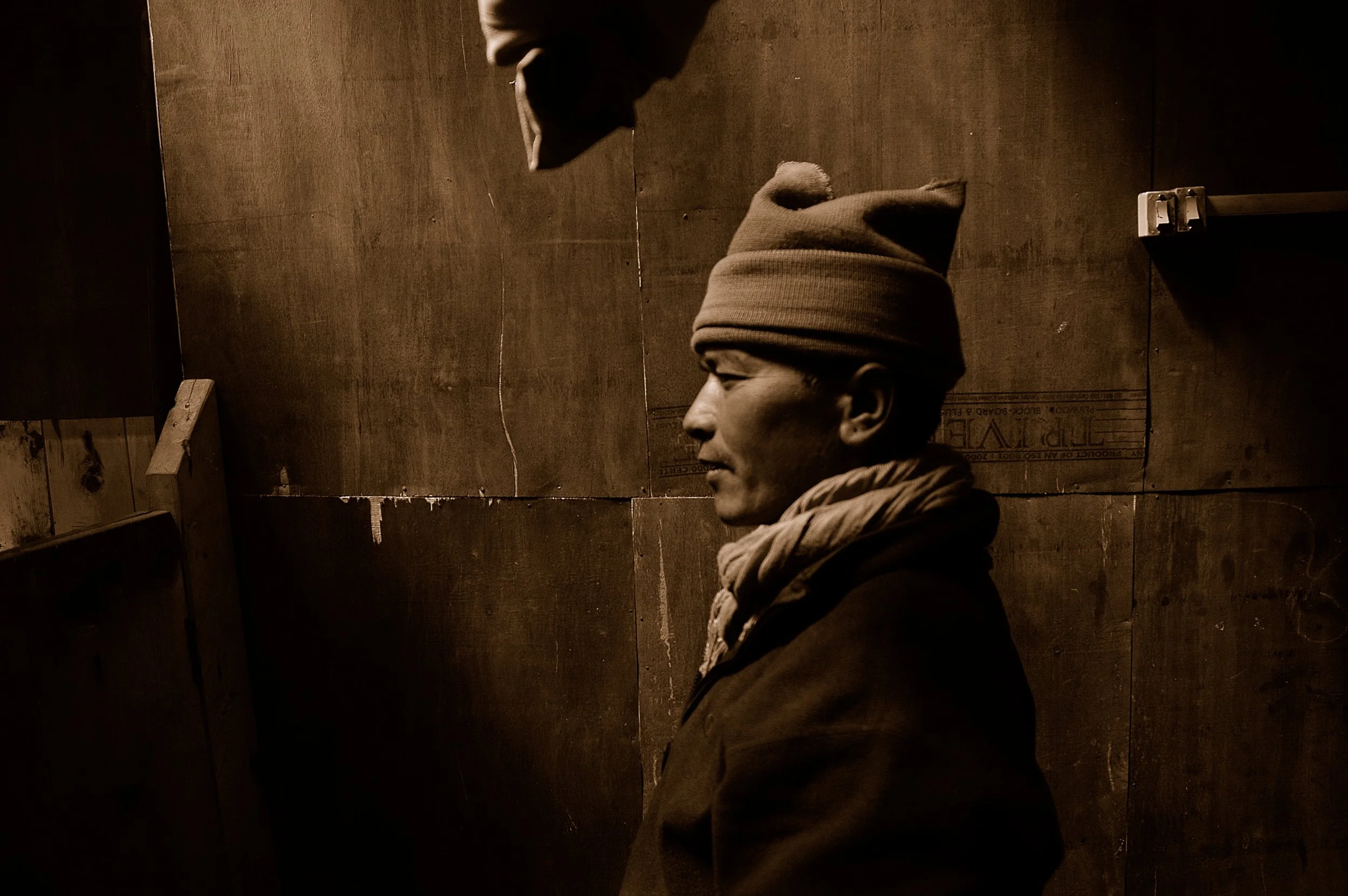 A side profile of a person wearing a beanie hat and a scarf, smiling with eyes closed, standing against a dark wooden background.