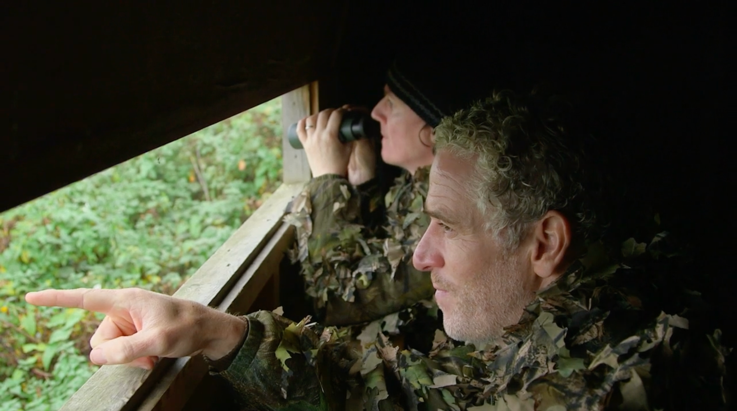 Two people in camouflage clothing looking out a small opening, with one using binoculars and the other pointing outside.