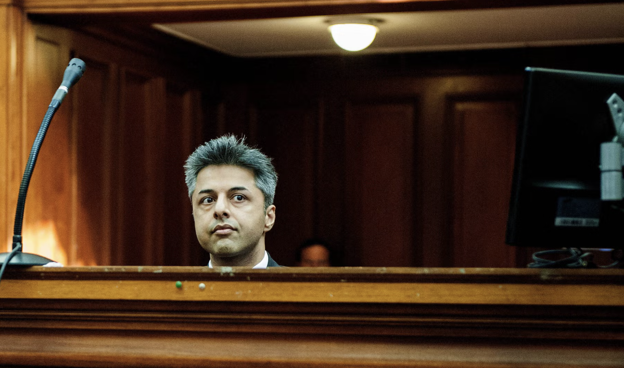 A man sitting in a courtroom or tribunal setting, looking serious and attentive, with a wooden panel in front of him and a monitor to his right.