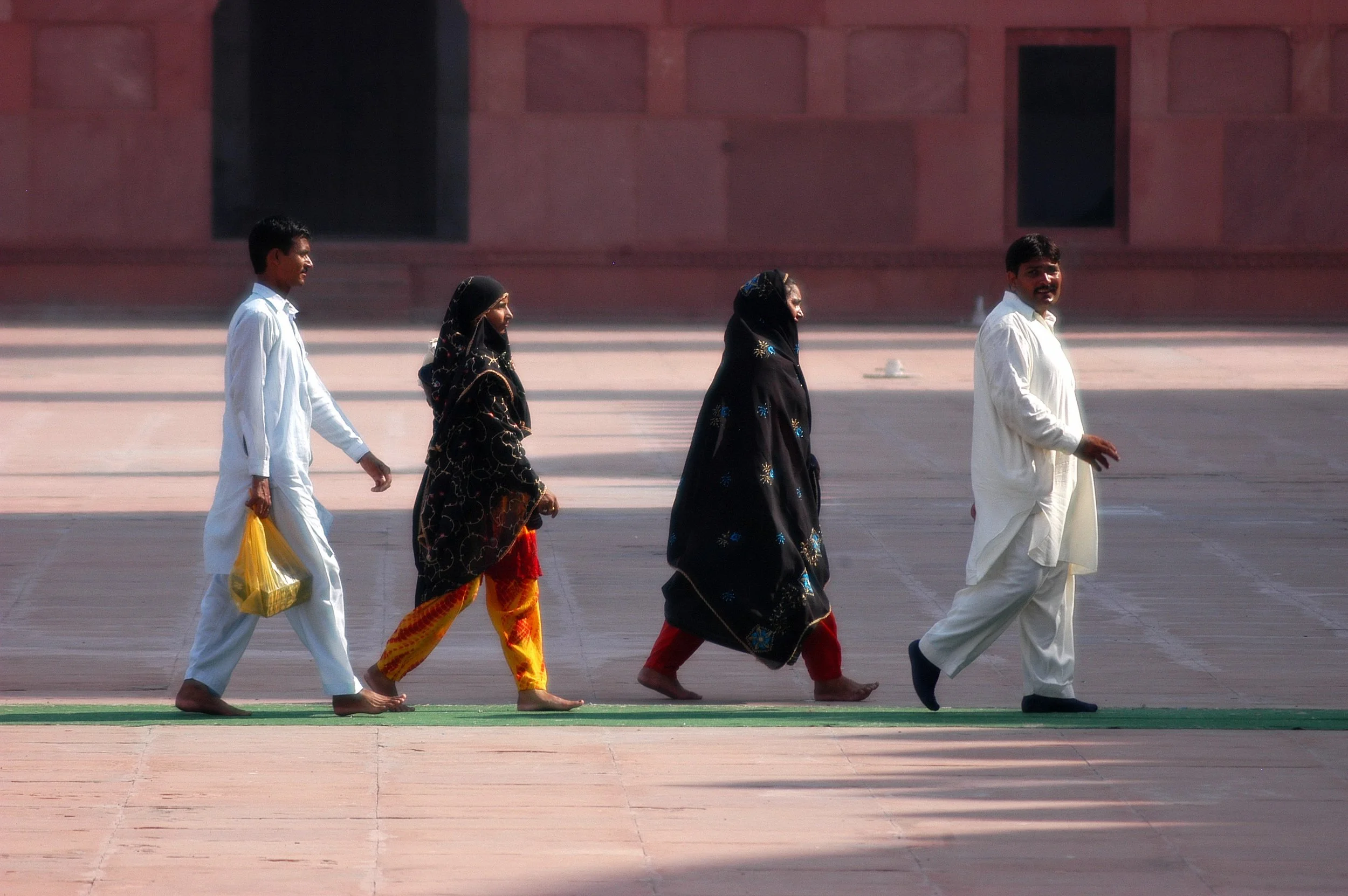 Five people, two men and three women, are walking barefoot on a large open paved area, possibly a historical or religious site. The men are dressed in traditional Indian white kurtas, while the women are wearing colorful, patterned sarees or shawls. 