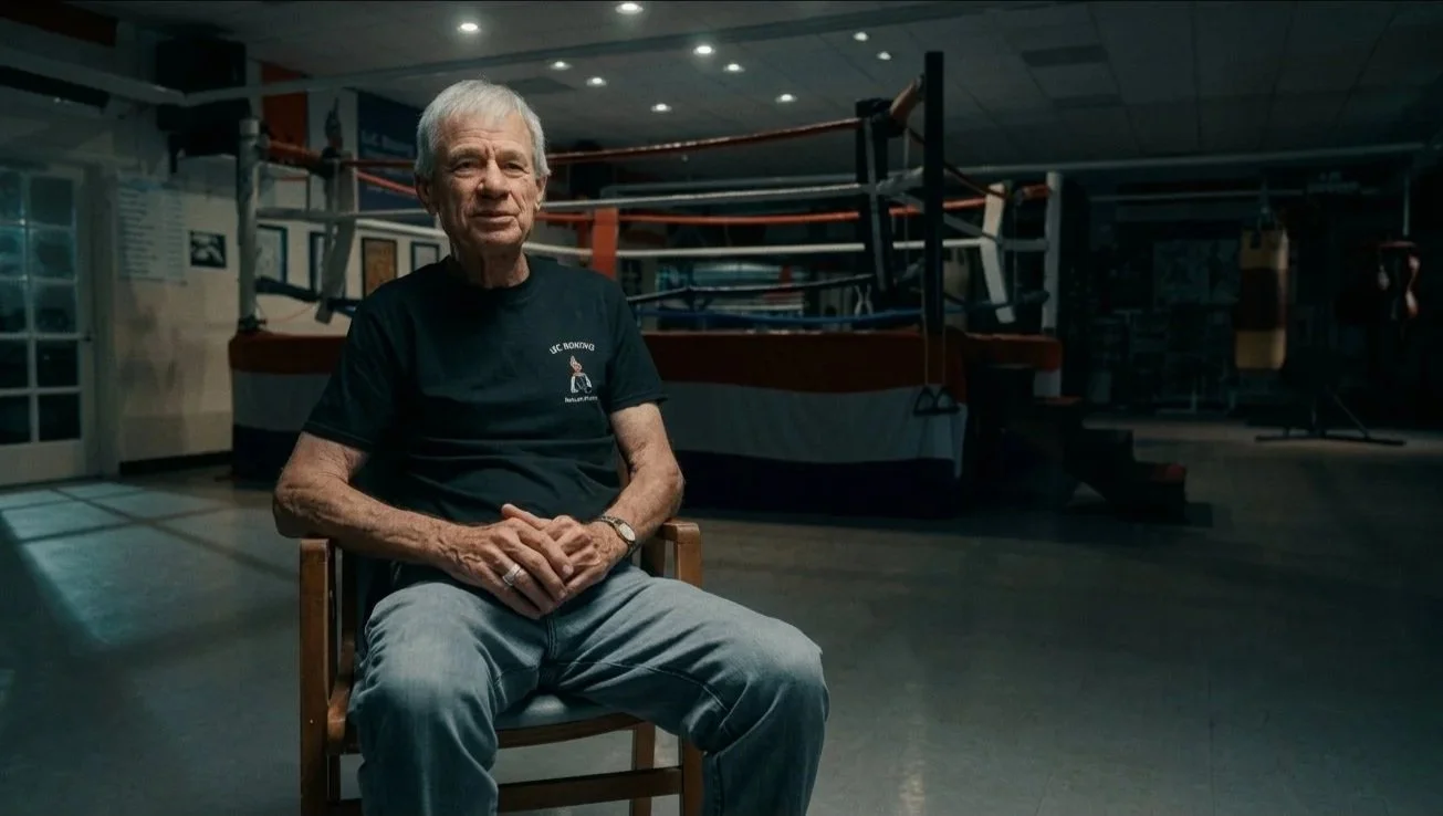An elderly man with white hair and wearing a black T-shirt sits on a chair inside a gym with a boxing ring in the background.
