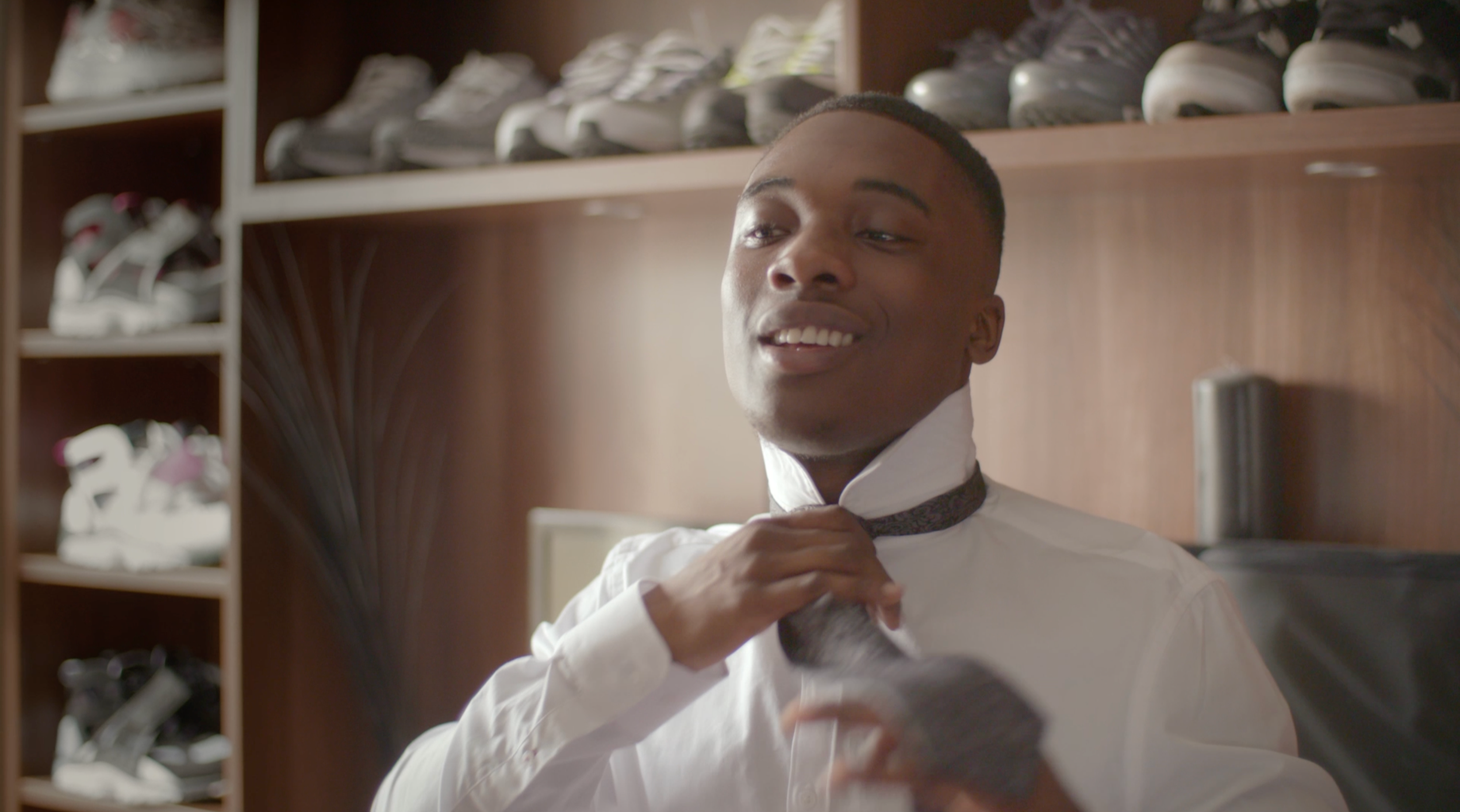 Young man in a white shirt adjusting his tie in front of a mirror, with shelves of sneakers in the background.