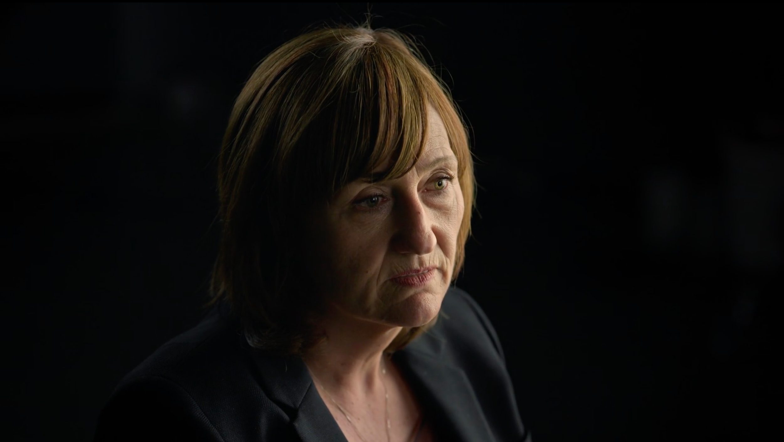 A middle-aged woman with reddish-brown hair looking contemplative against a black background.