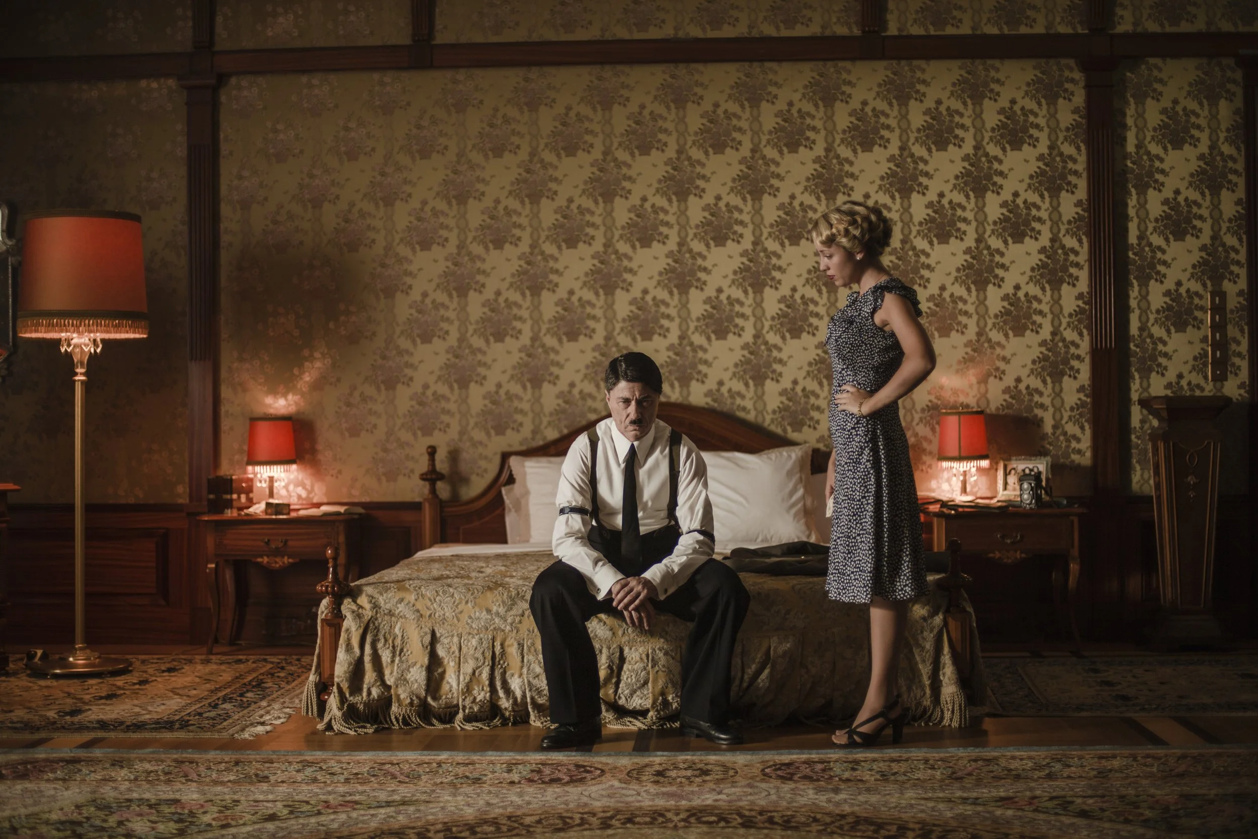 A man in a white shirt with rolled-up sleeves and black tie sitting on a bed, with a woman in a dark patterned dress standing beside him, in a vintage-style room with floral wallpaper, wooden furniture, and multiple lamps.