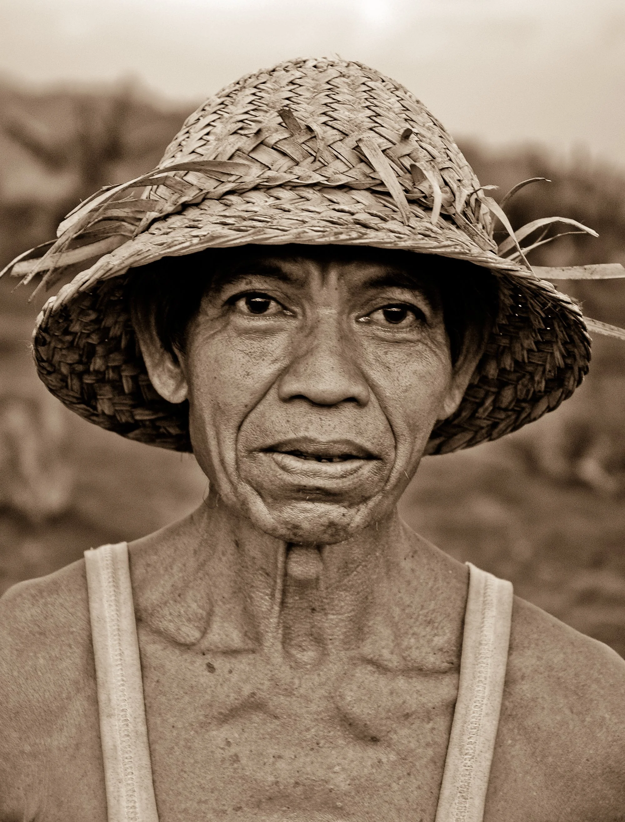 A close-up portrait of an elderly person wearing a woven straw hat, with a background of a field.