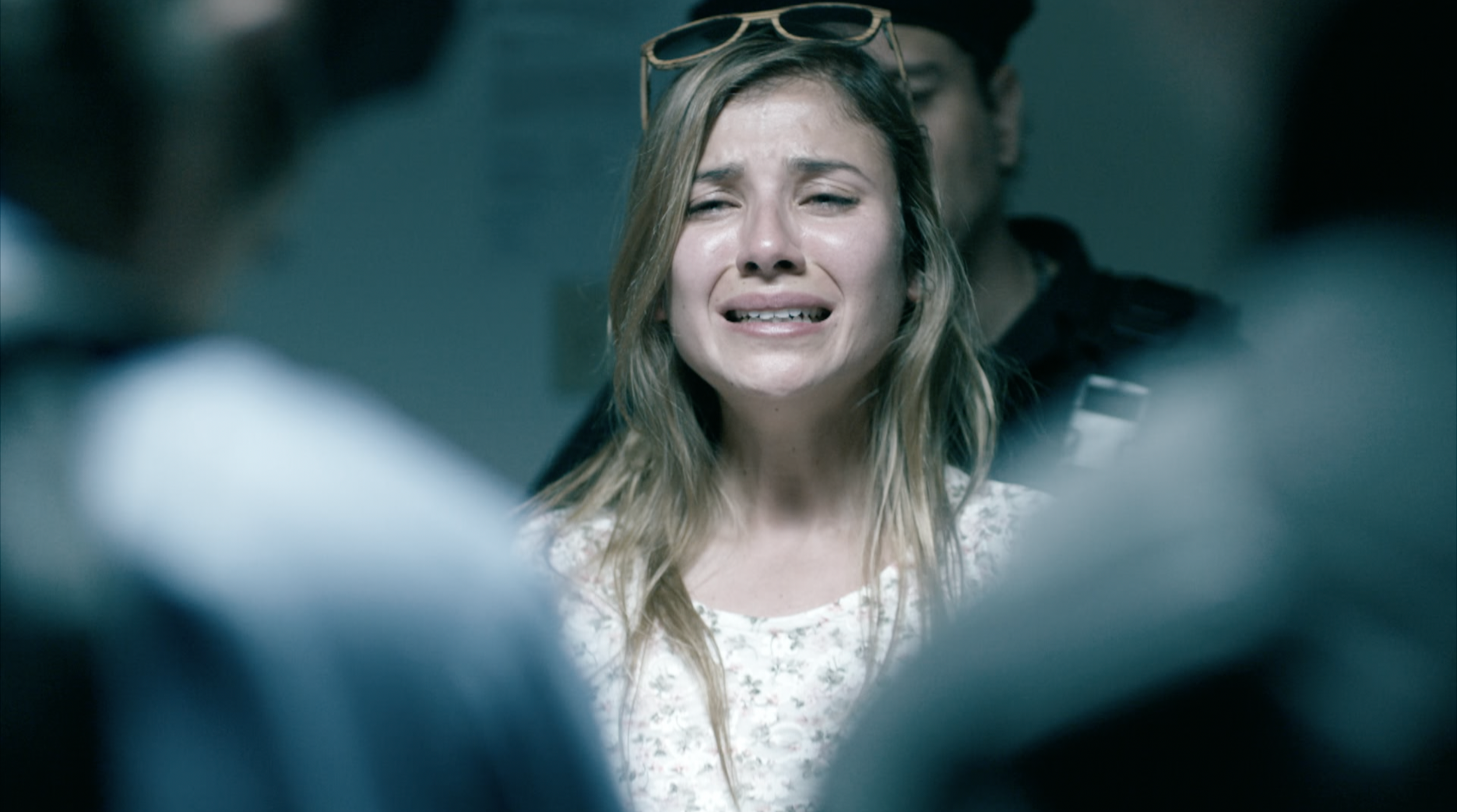 A woman with long, wavy blonde hair, wearing a white floral dress and sunglasses on her head, appears to be upset or crying.