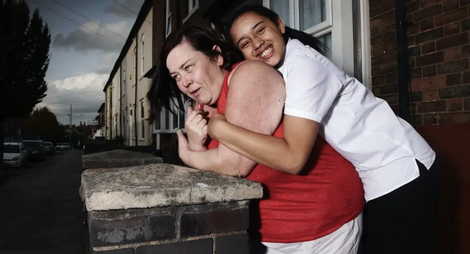 Two women hugging on a sidewalk, one with darker skin and black hair, smiling, in a white shirt, the other with lighter skin, dark hair, unhappy expression, in a red dress, near brick buildings and cars parked on the street.