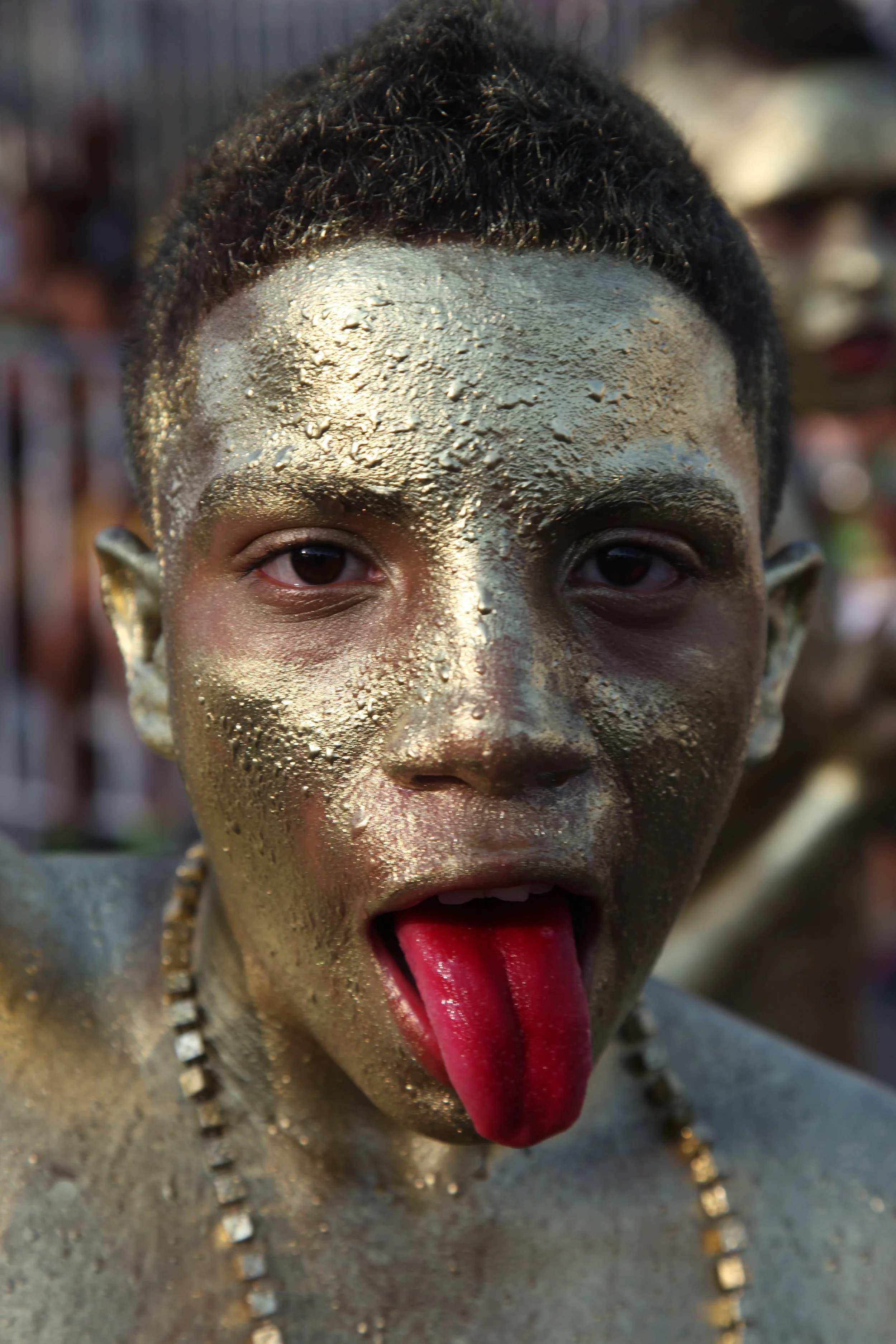 A person with black, curly hair and gold paint on their face, sticking out their tongue with red paint on it. They are wearing necklaces and have earrings, with a blurred background.