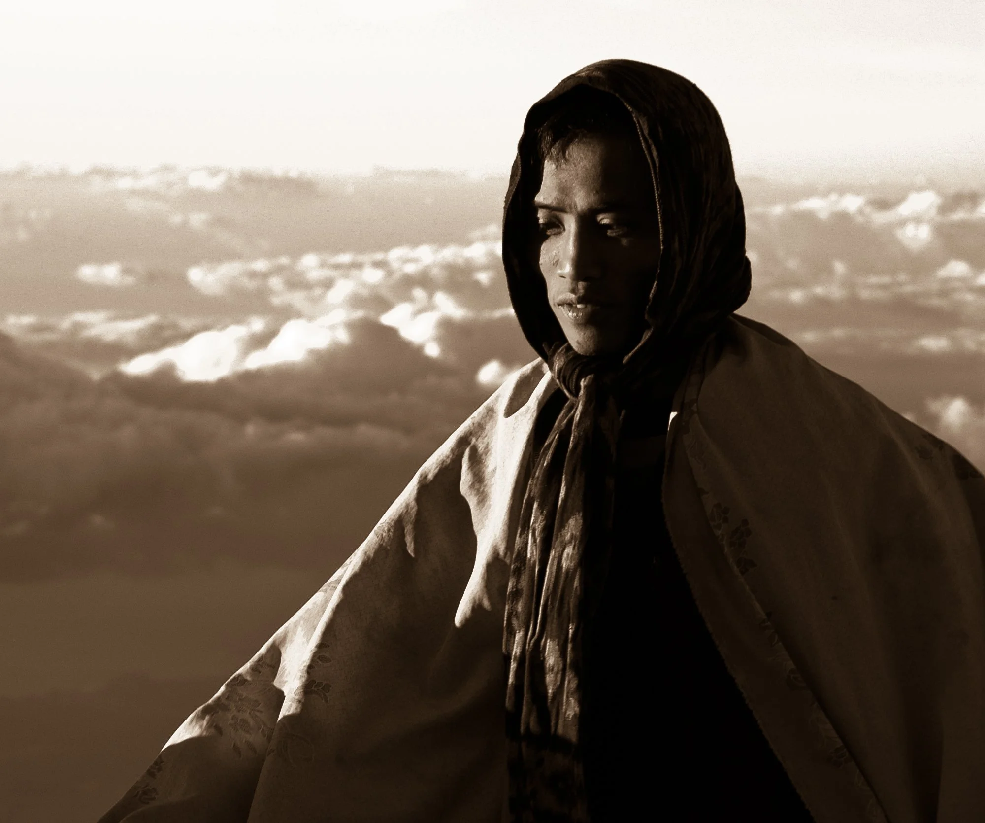 A woman with dark skin wearing a headscarf, looking down, against a cloudy sky background in black and white.