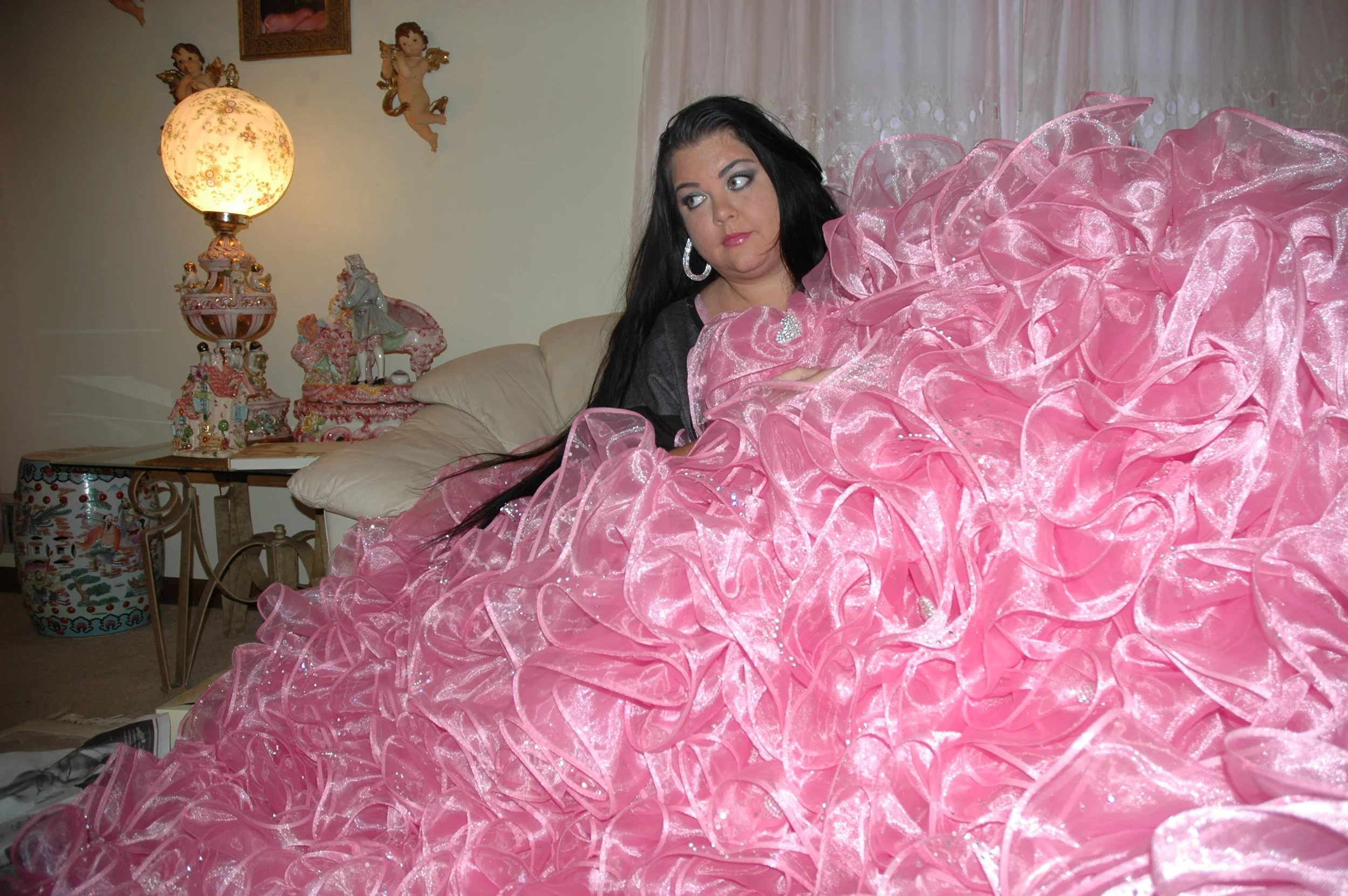 A woman with long dark hair and hoop earrings is lying on a bed with a large pink ruffled dress covering her. The room features a lamp with a floral design, figurines, and wall decorations including cherubs and a small painting.