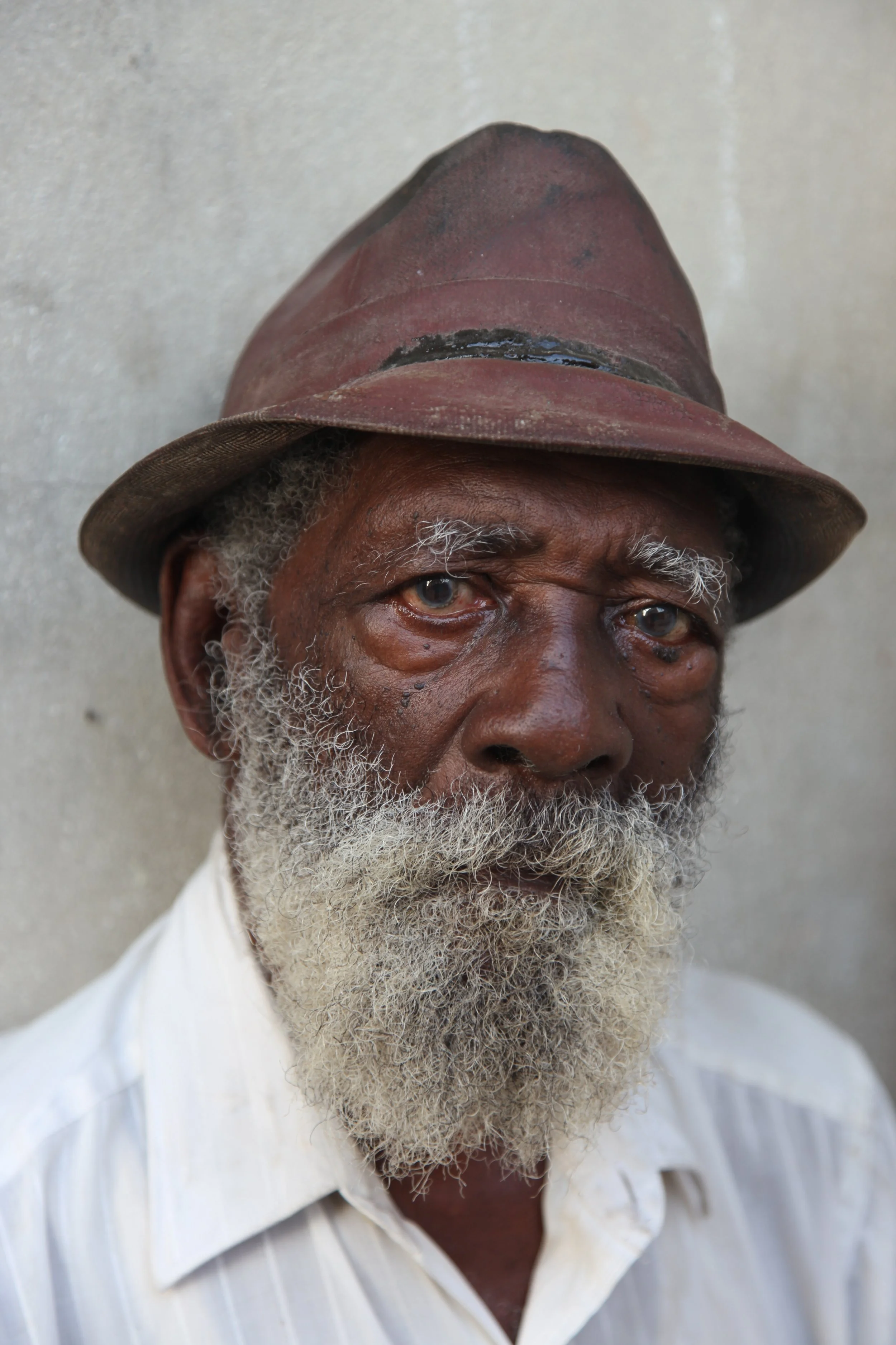 Close-up of an elderly man with a white and gray beard, wearing a weathered brown fedora hat and a white, striped collared shirt, against a plain neutral background.