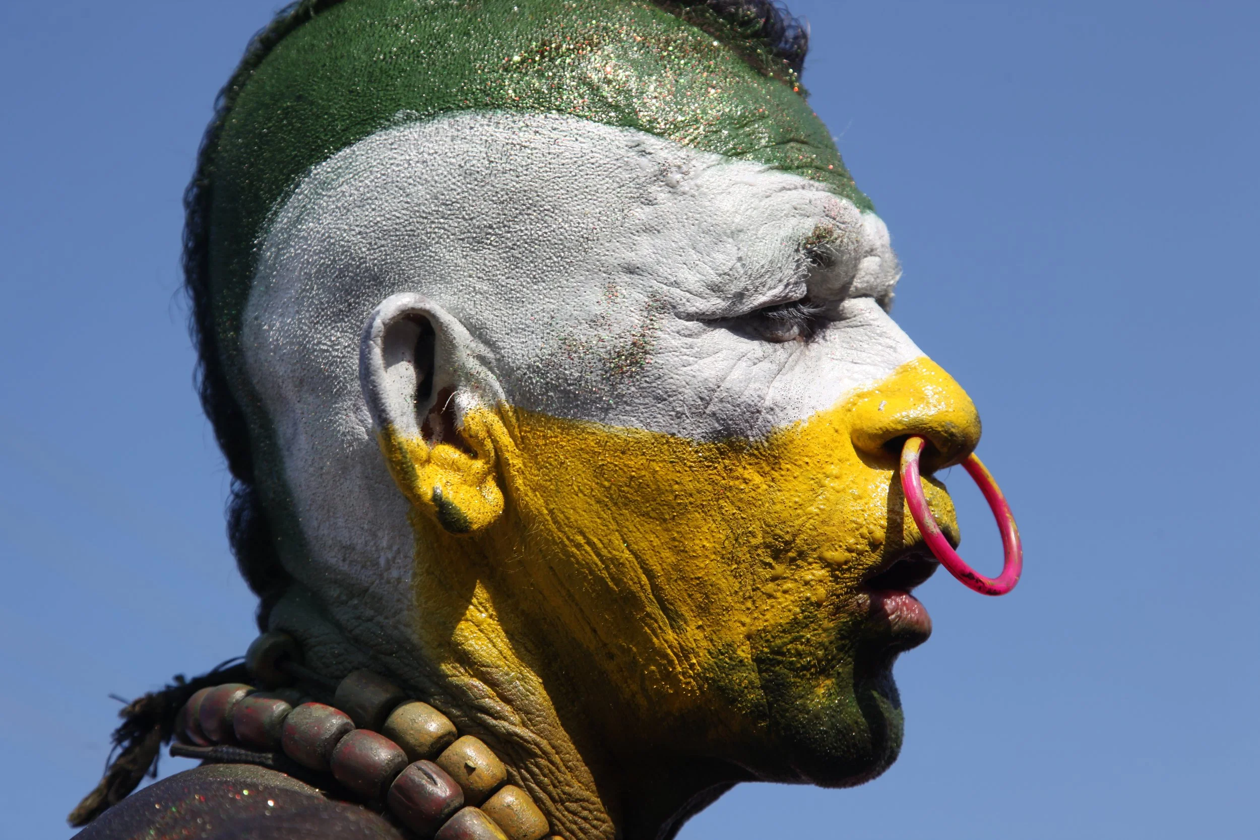 Close-up of a person with their face painted in green, white, and yellow colors, wearing jewelry, and with a pink nose ring against a clear blue sky.