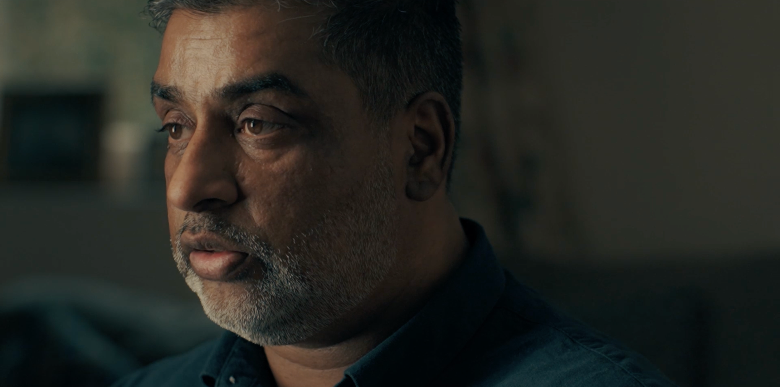 Close-up of a middle-aged man with short, dark hair and a gray beard, looking to the side, in a dim indoor setting.
