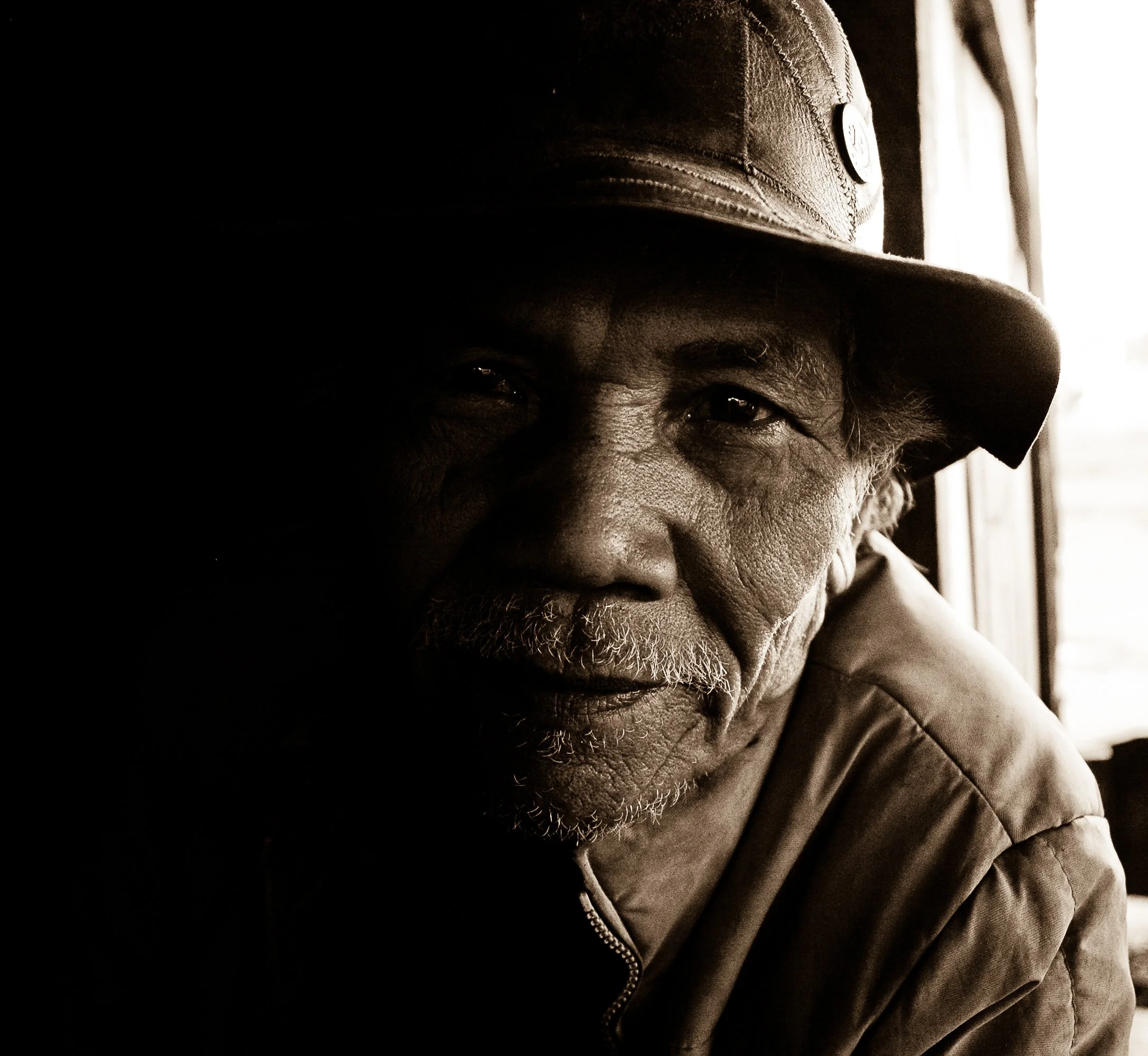 Close-up portrait of an African American man wearing a baseball cap and jacket, smiling with his eyes. The image uses a sepia filter with strong shadows.
