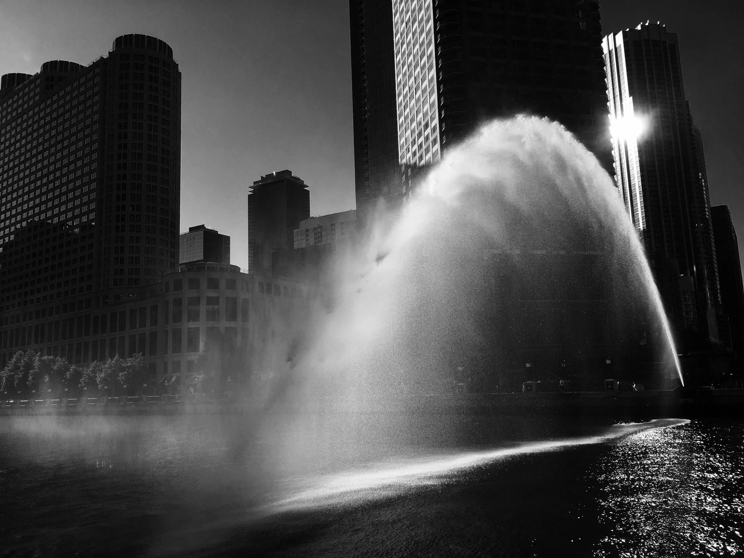 A black and white photo of tall skyscrapers in a city with a water fountain in the foreground, with water spraying up in front of the buildings.